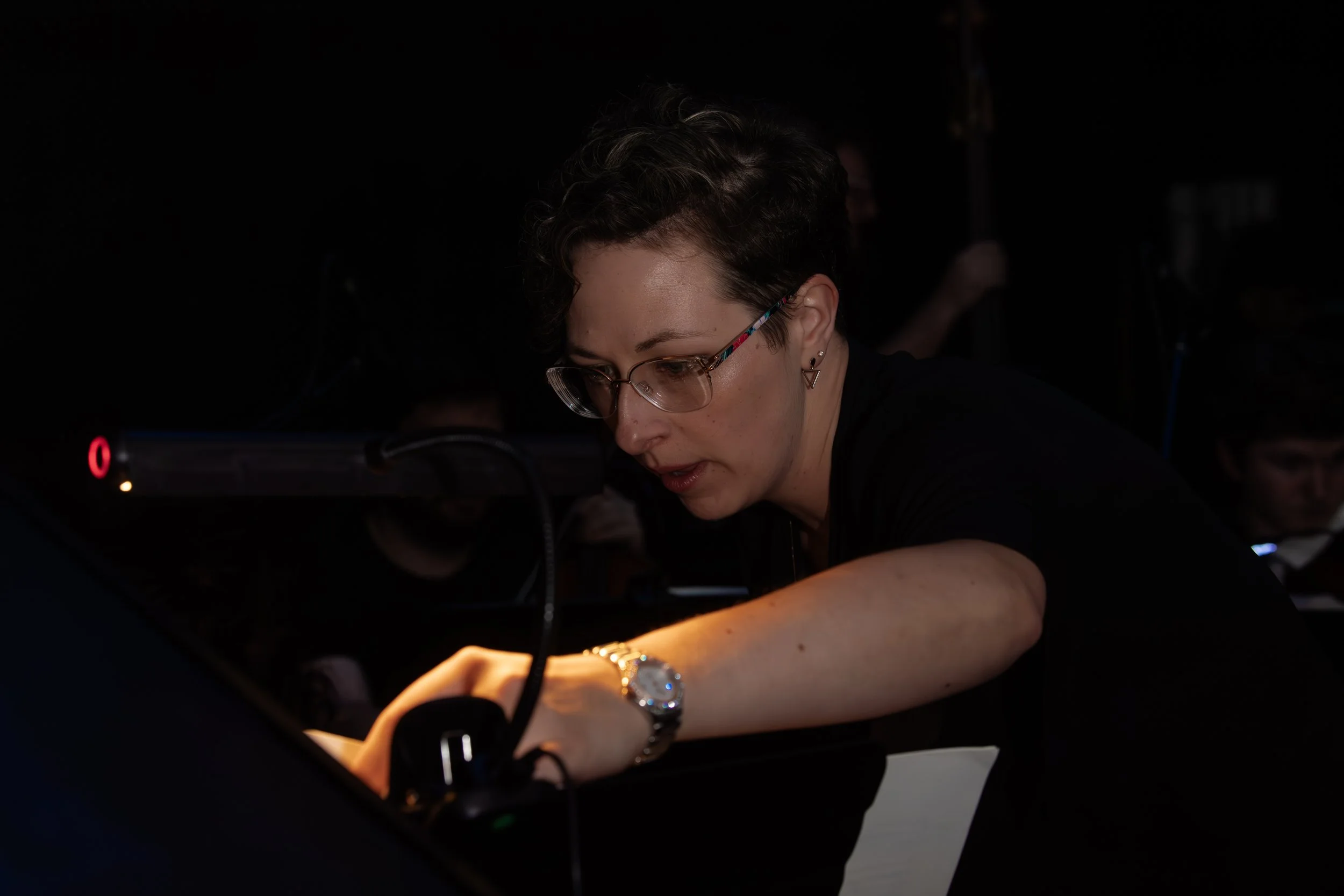 Woman with short dark hair, glasses, and earrings working on a computer in a dark room with focused lighting.