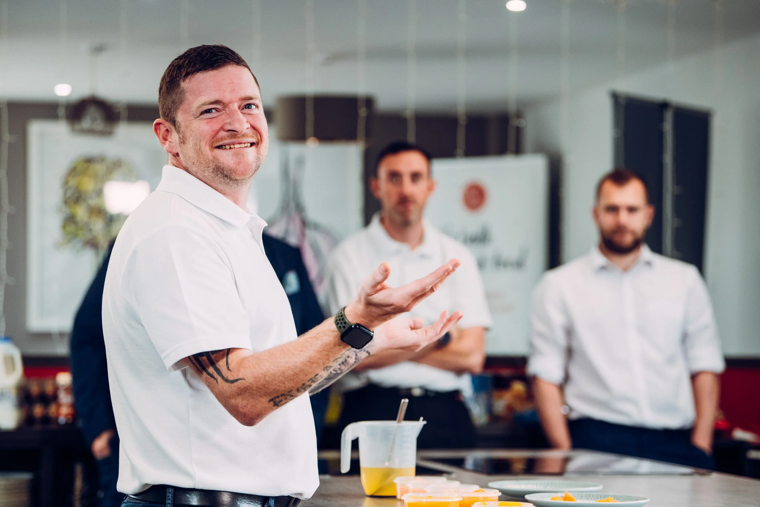 Man with short brown hair, beard, and tattoos on his arm, smiling and gesturing with his hand in a kitchen or cooking class setting, with three other men in white shirts standing in the background.