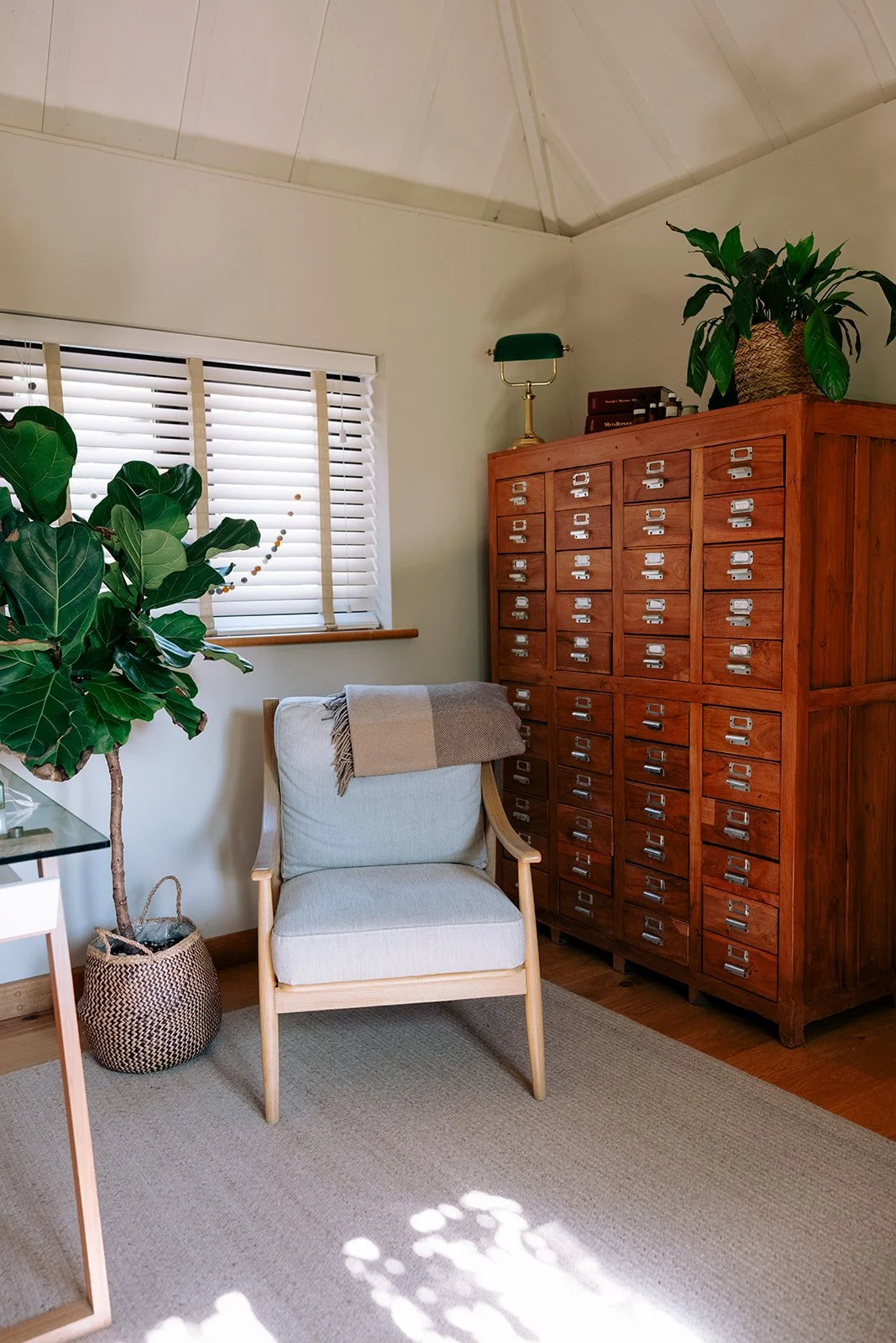 A woman in a white dress seated in a wooden armchair, reading a book in a cozy, well-decorated home office or living space with a computer on a white desk, plants, wall art, and a window with blinds, sunlight casting shadows on the floor.