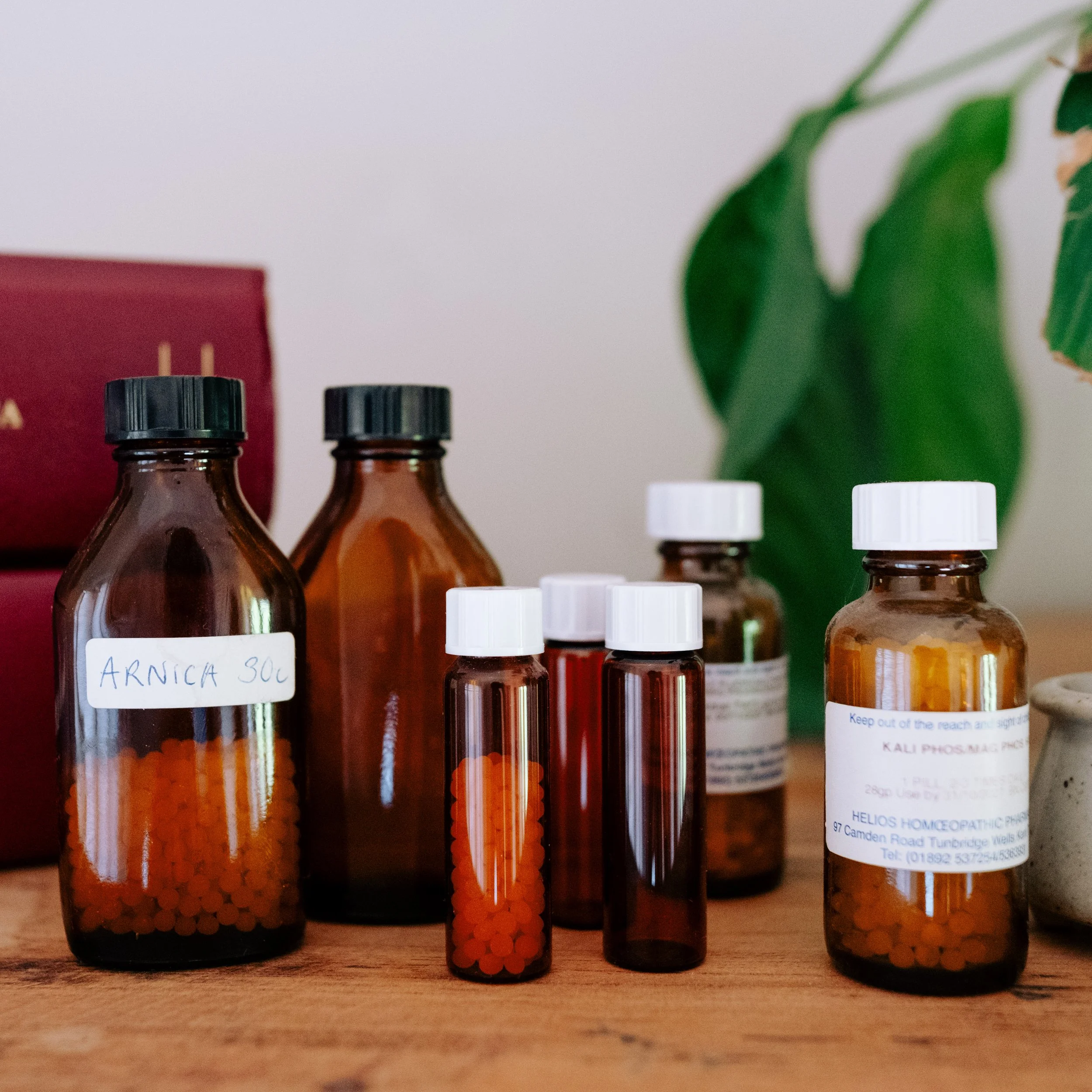 Brown glass bottles and small vials containing orange homeopathic pills on a wooden surface, with a plant in the background.