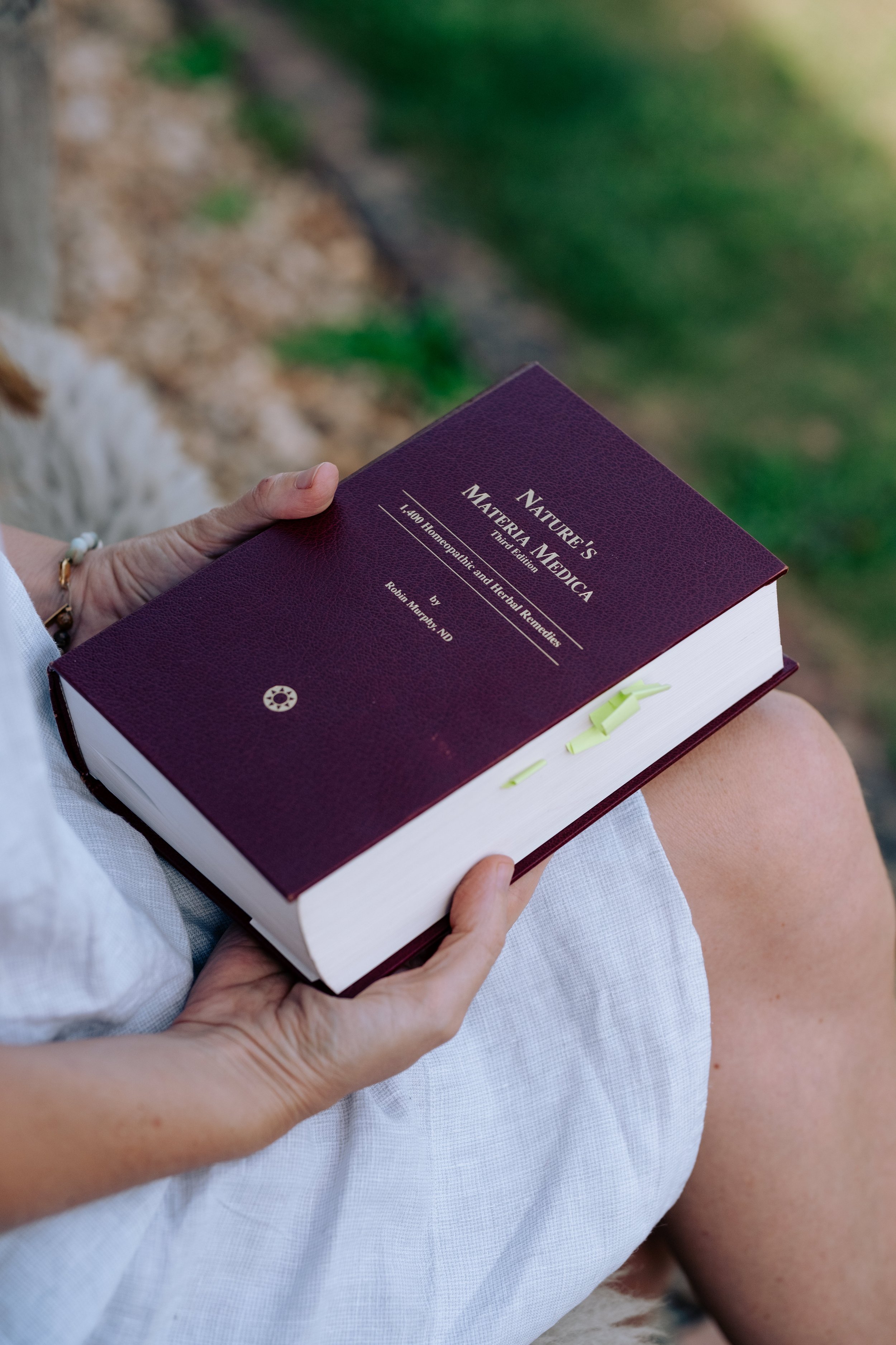 Person holding a large hardcover book titled 'NATURE'S MATERIALS MEDICA' outdoors, with green grass and ground in the background.