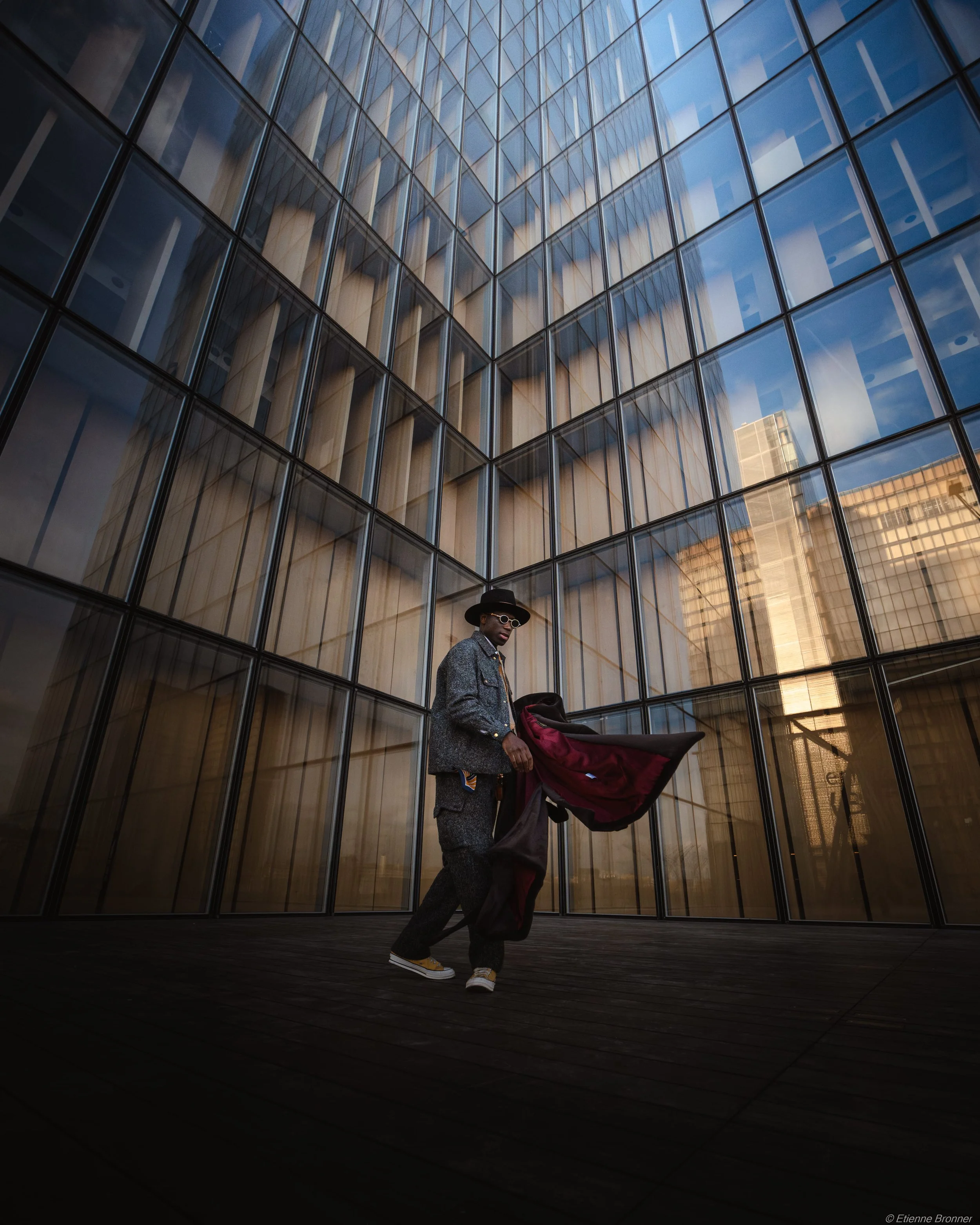 Une personne habillée élégamment portant un manteau gris, un chapeau noir et des lunettes, se tenant devant un grand bâtiment en verre moderne au coucher du soleil.
