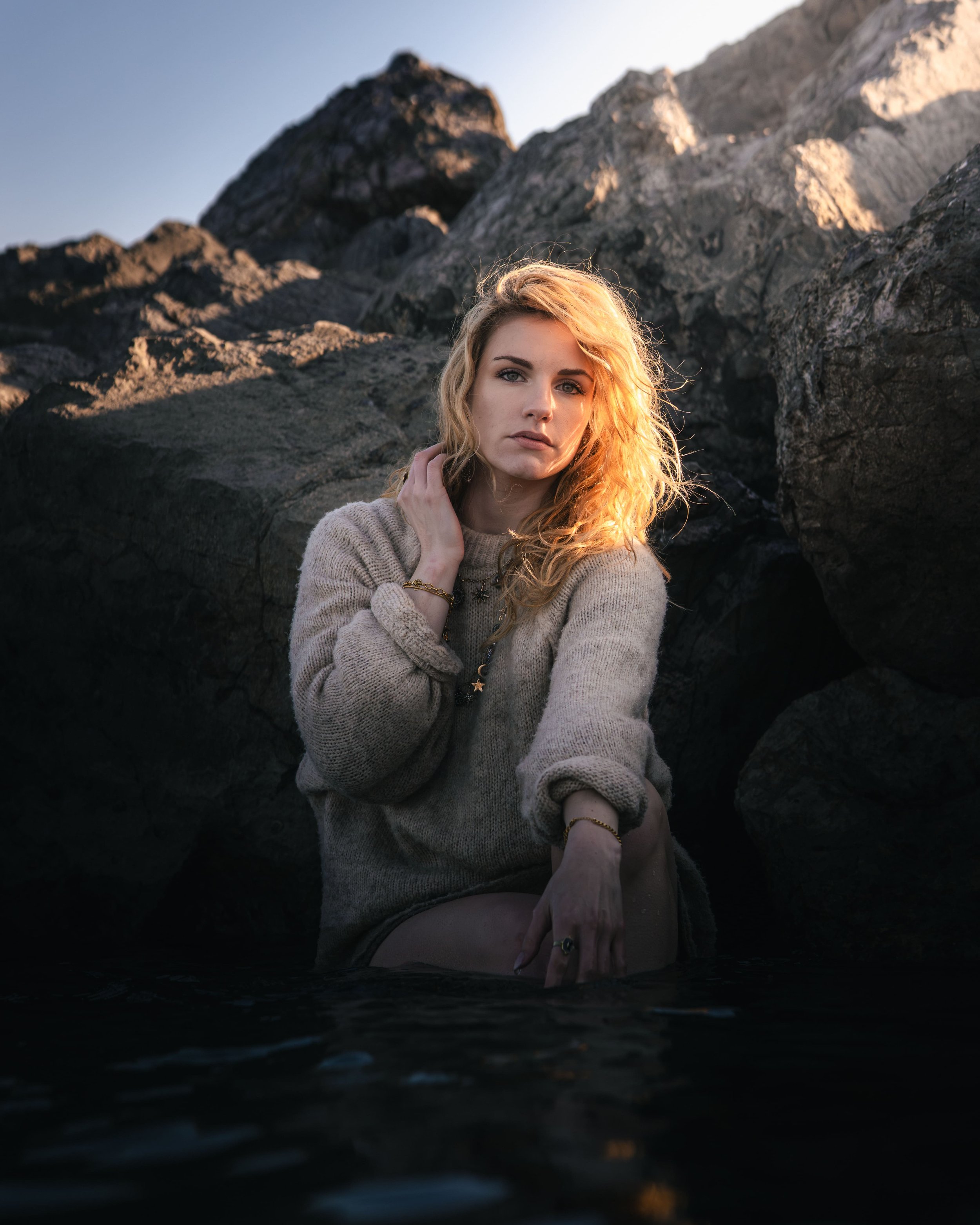 Jeune femme aux cheveux bouclés blonds, portant un pull beige, assise dans l'eau près de rochers, regardant vers la caméra au coucher de soleil.