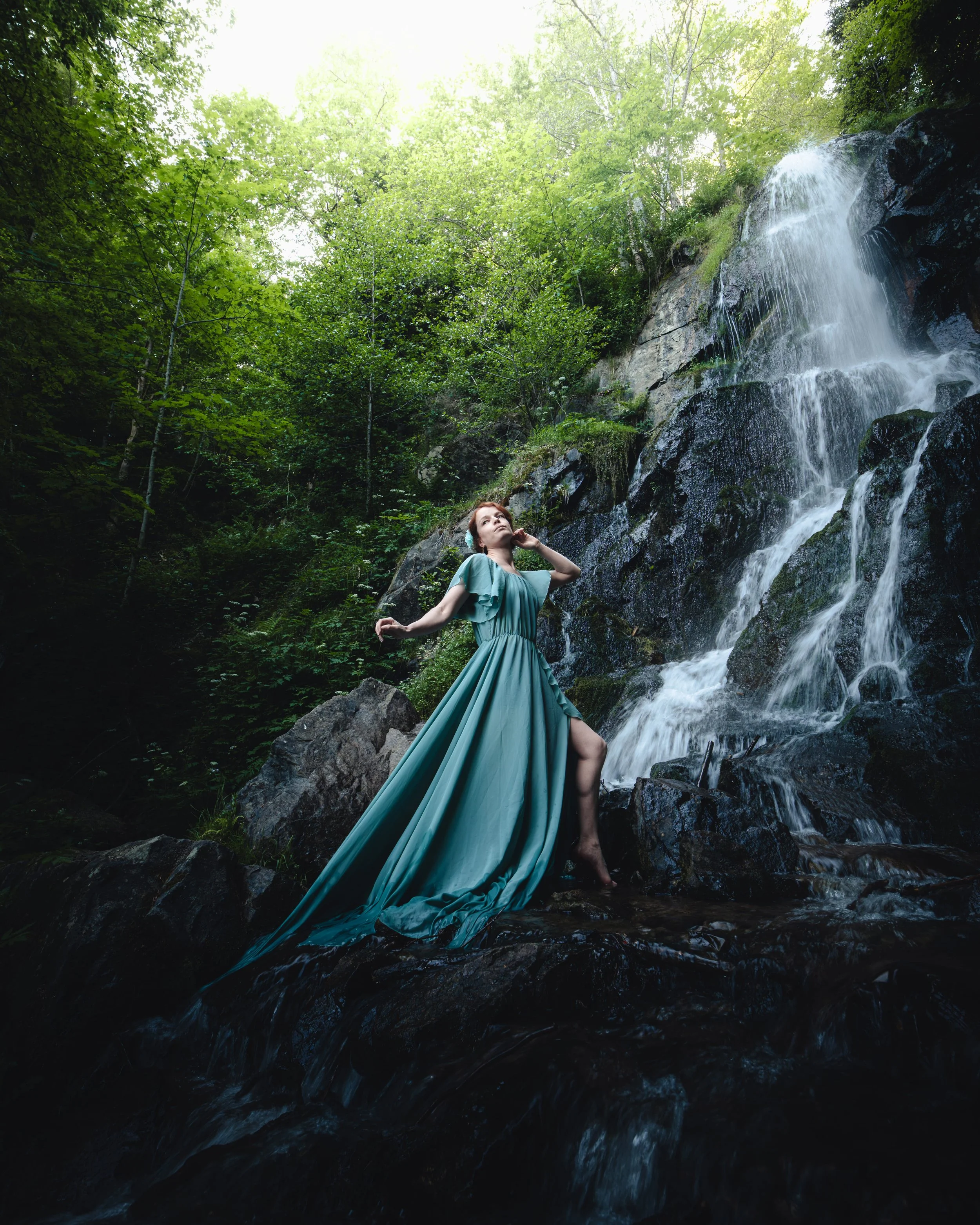 Portrait d'une femme en robe longue turquoise posant sur des rochers près de la cascade du Hohwald en Alsace, dans une forêt verdoyante, sous un ciel lumineux.