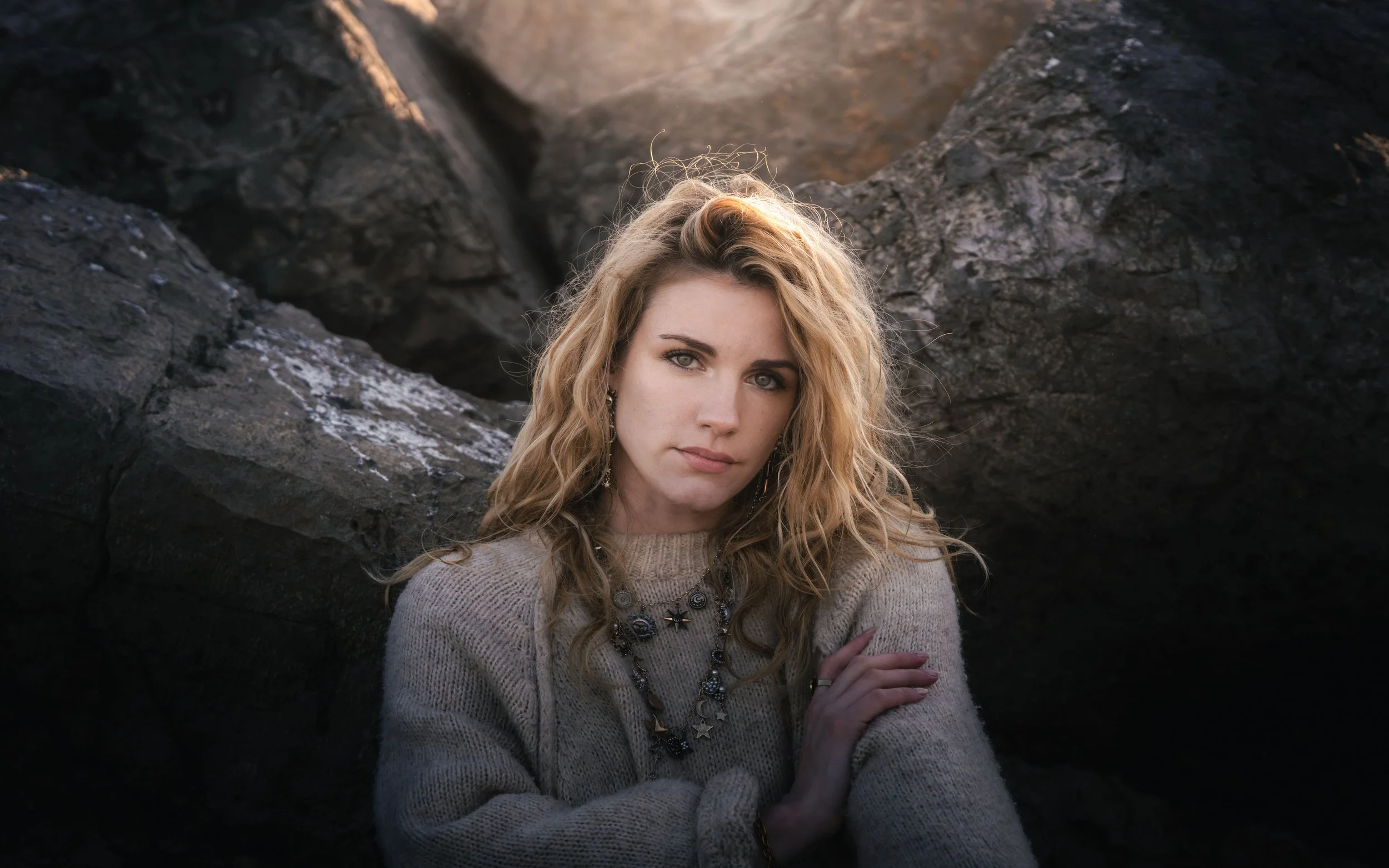Femme aux cheveux bouclés blonds portant un pull en laine beige et un collier, posant devant des rochers.