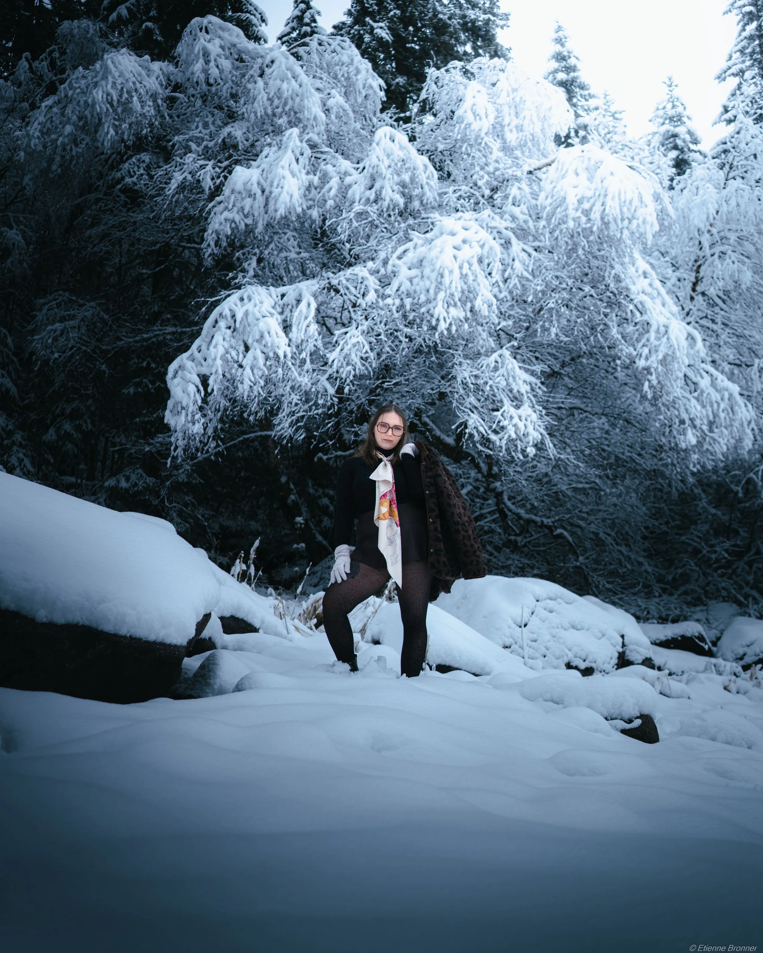 Portrait d'une femme portant des vêtements élégants pose dans un paysage enneigé avec de grands arbres couverts de neige en arrière-plan, dans les Vosges.