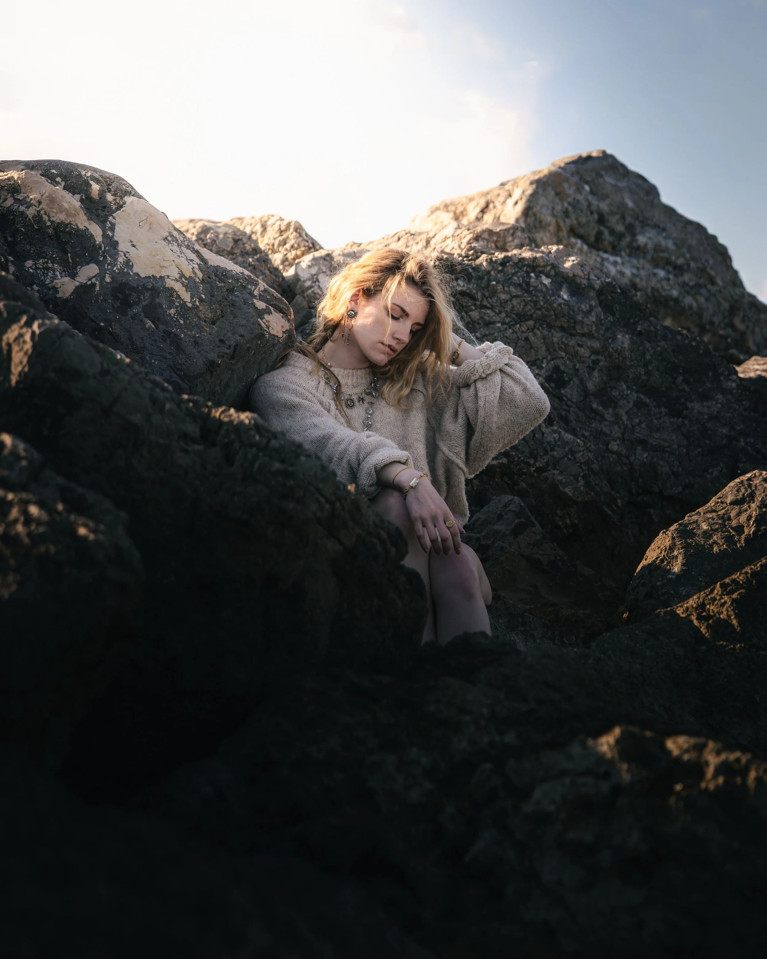 Une femme blonde assise sur des rochers, portant un pull beige, dans un paysage rocheux sous un ciel clair.