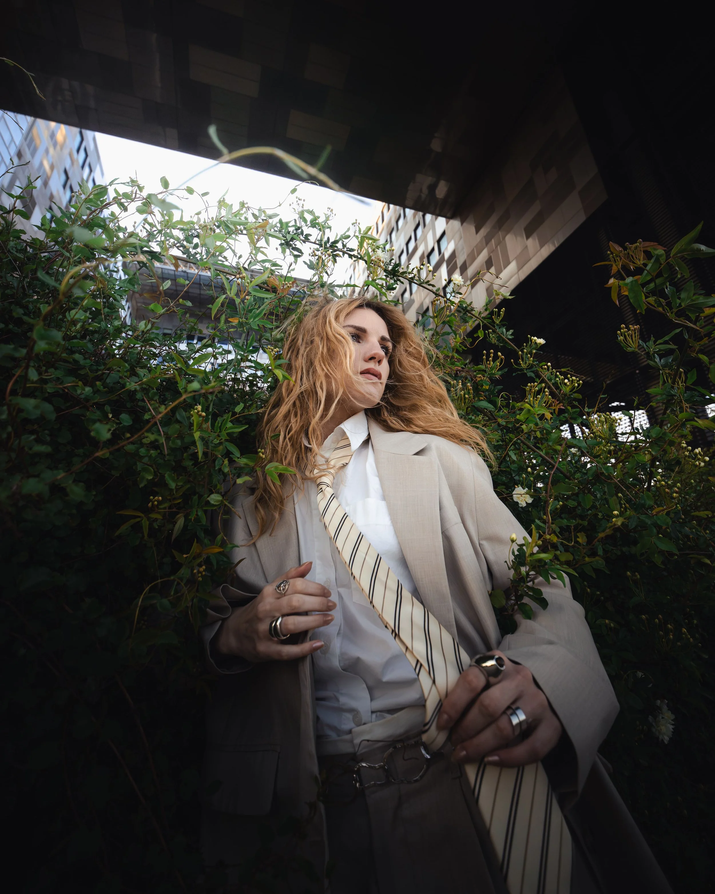 Femme avec des cheveux bouclés orange portant un costume beige et une chemise blanche, debout devant une haie verte. Vêtue de plusieurs bagues et d'une cravate rayée, regardant vers la droite, dans un environnement urbain.