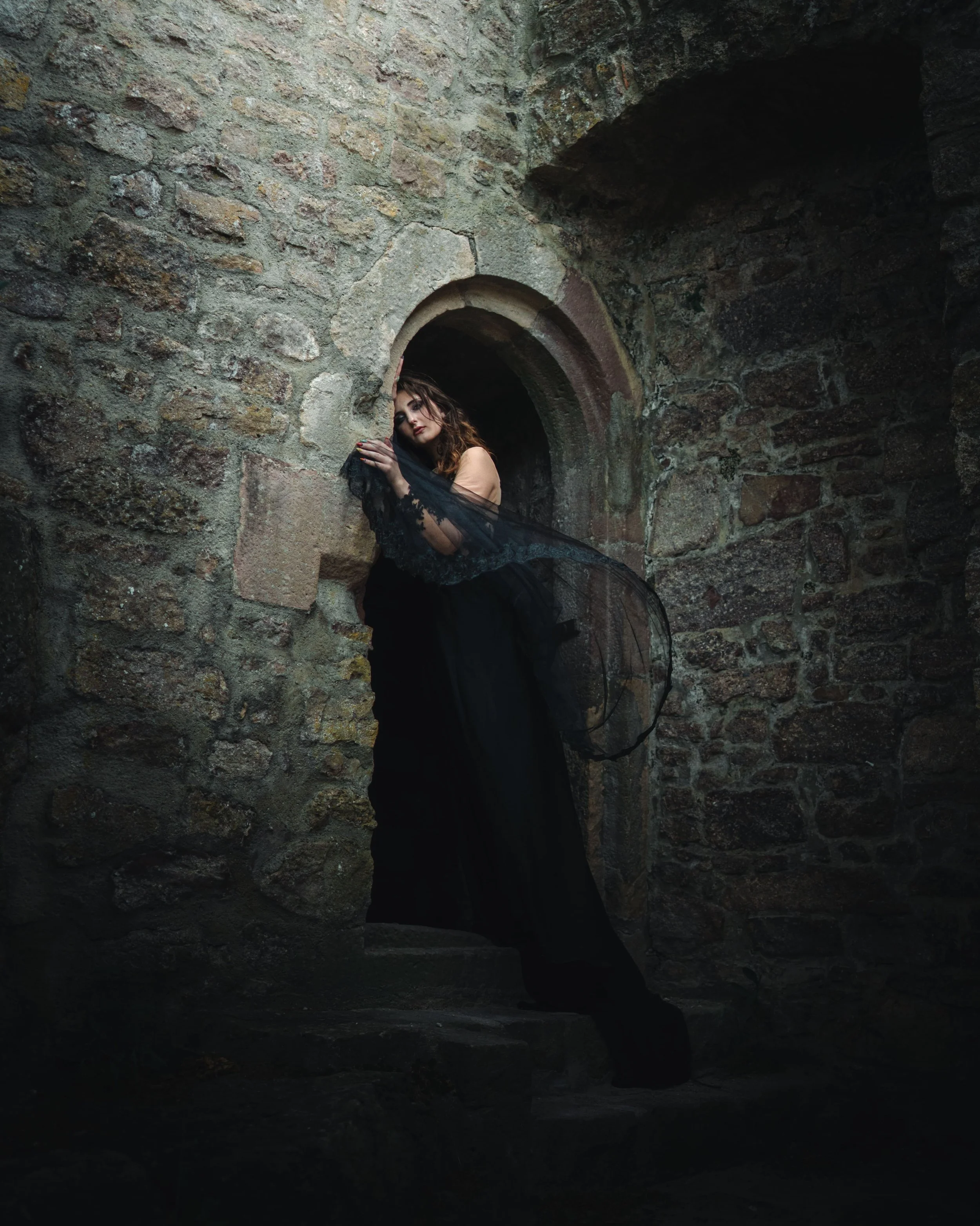 Portrait d'une femme avec des cheveux bruns, vêtue de noir, posant dans une fenêtre ovale en pierre dans un mur en pierre vieux. La scène a une atmosphère sombre et mystérieuse. Au château de Hohenbaden en Allemagne.