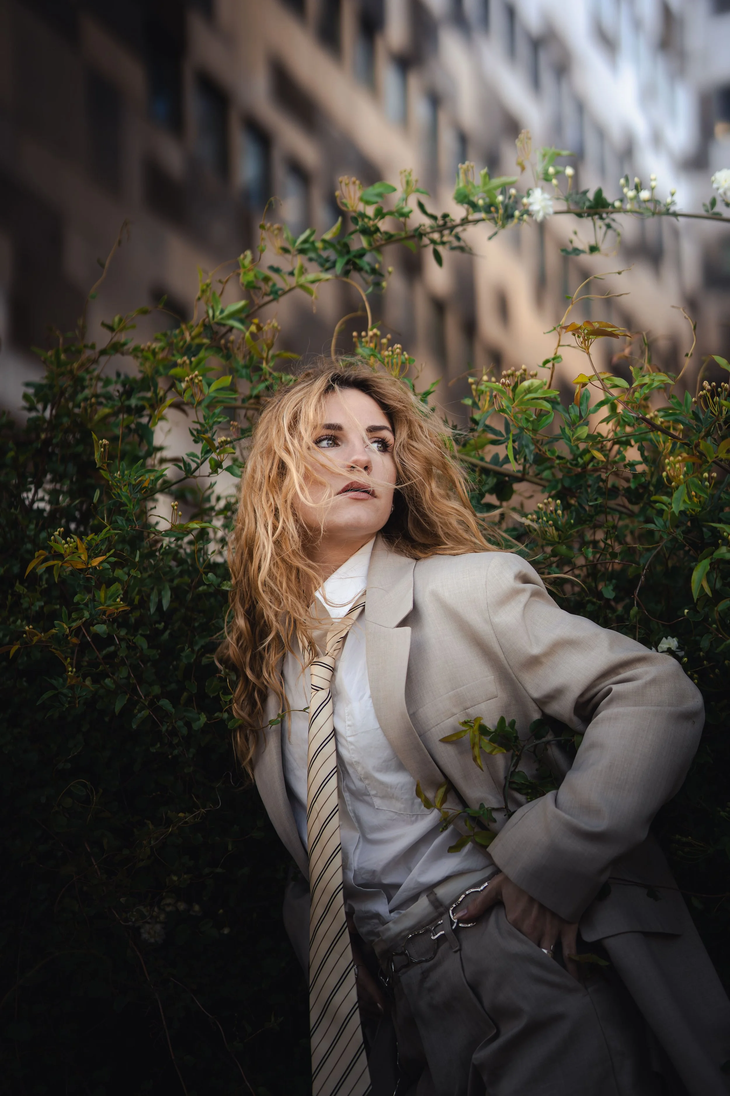 Une femme blonde habillée d'un costume beige, debout près de buissons verts, avec un bâtiment en arrière-plan.