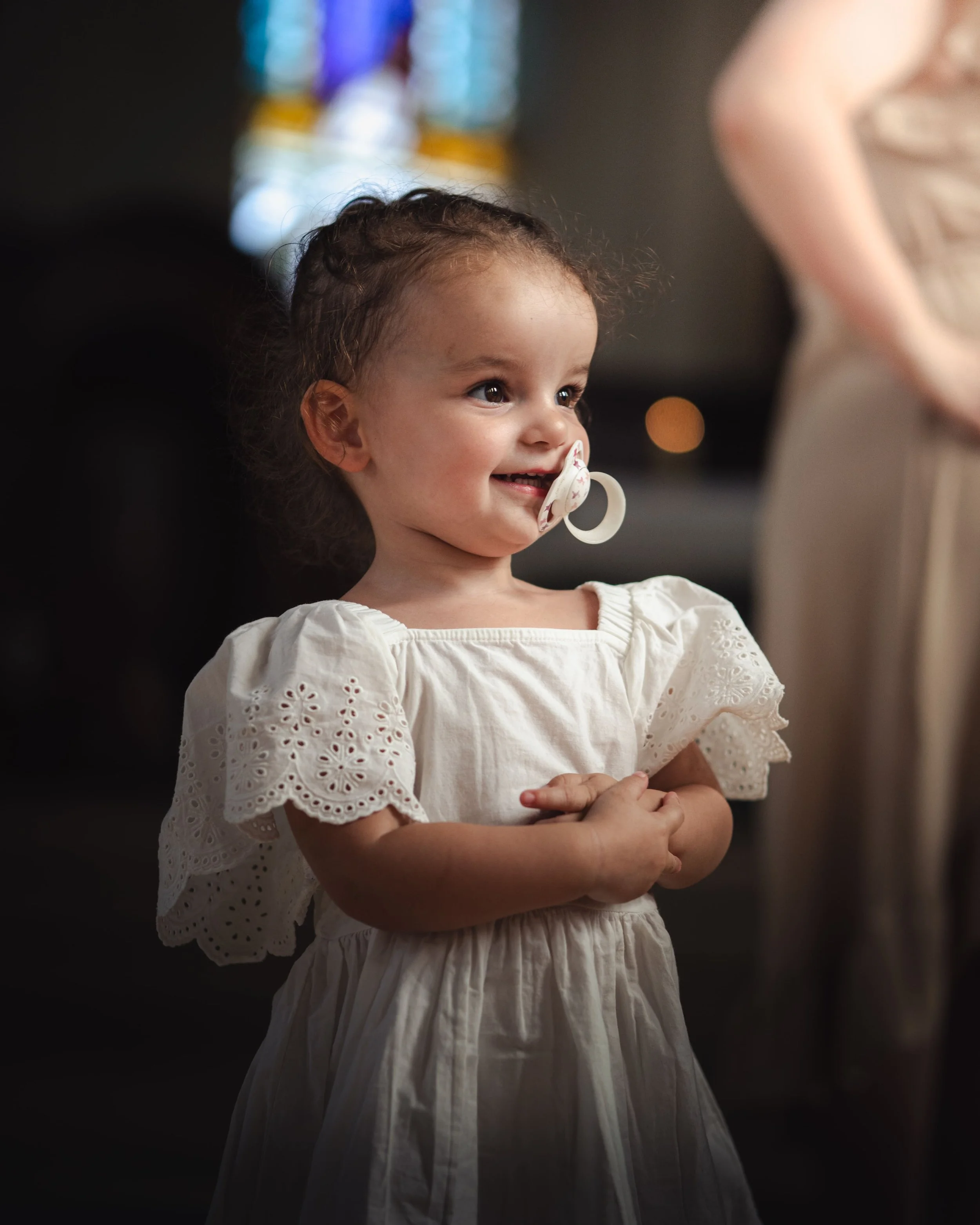 Portrait d'une jeune fille souriante portant une robe blanche en dentelle, avec une tétine dans la bouche, dans un environnement intérieur, lors d'un baptême.