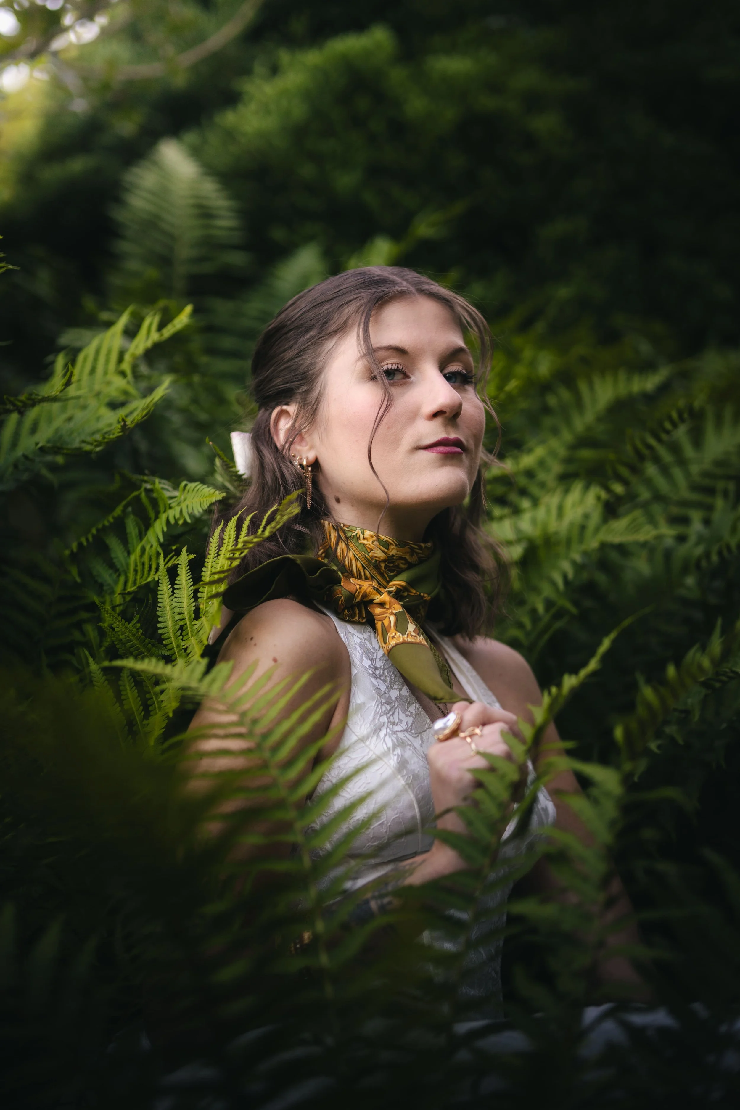 Une femme avec des cheveux bruns ondulés, portant un foulard en soie Hermès et une bague, se tient parmi des fougères vertes dans la forêt, regardant légèrement de côté.