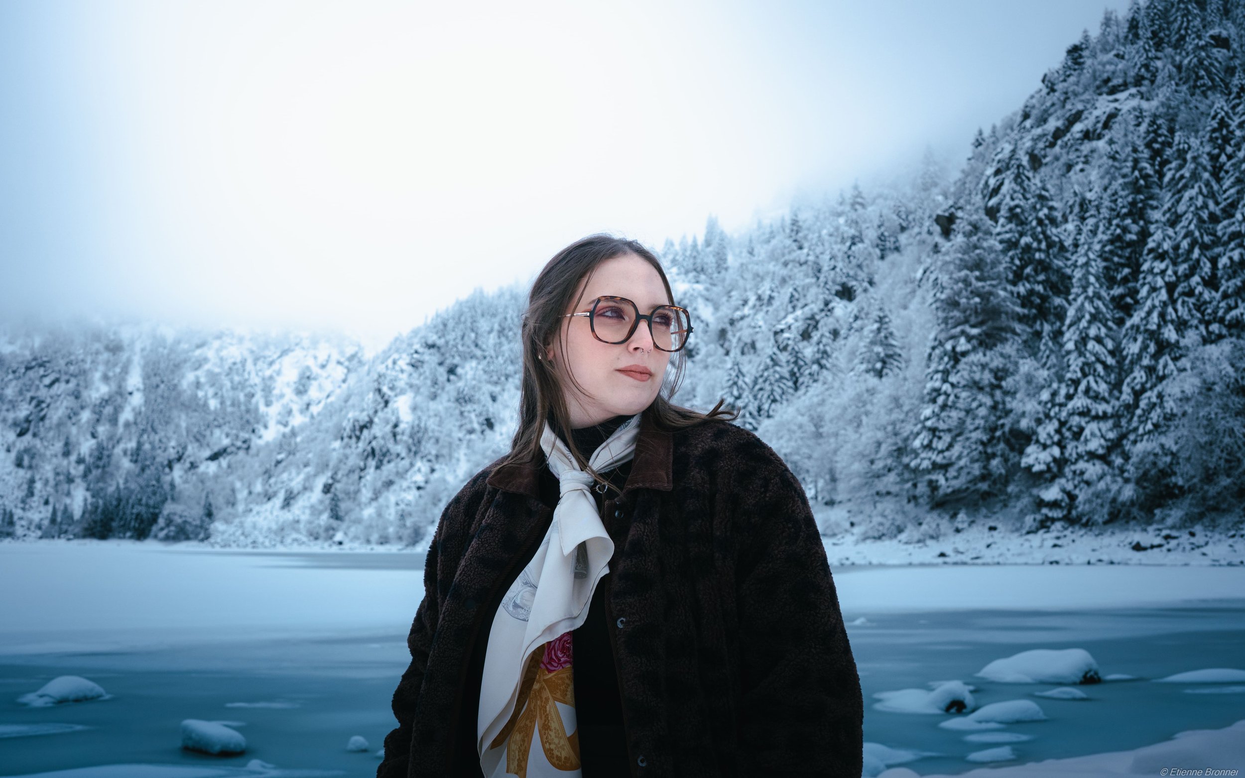 Portrait d'une femme portant des lunettes et un foulard blanc se tient près du lac blanc dans les Vosges avec des montagnes enneigées en arrière-plan.