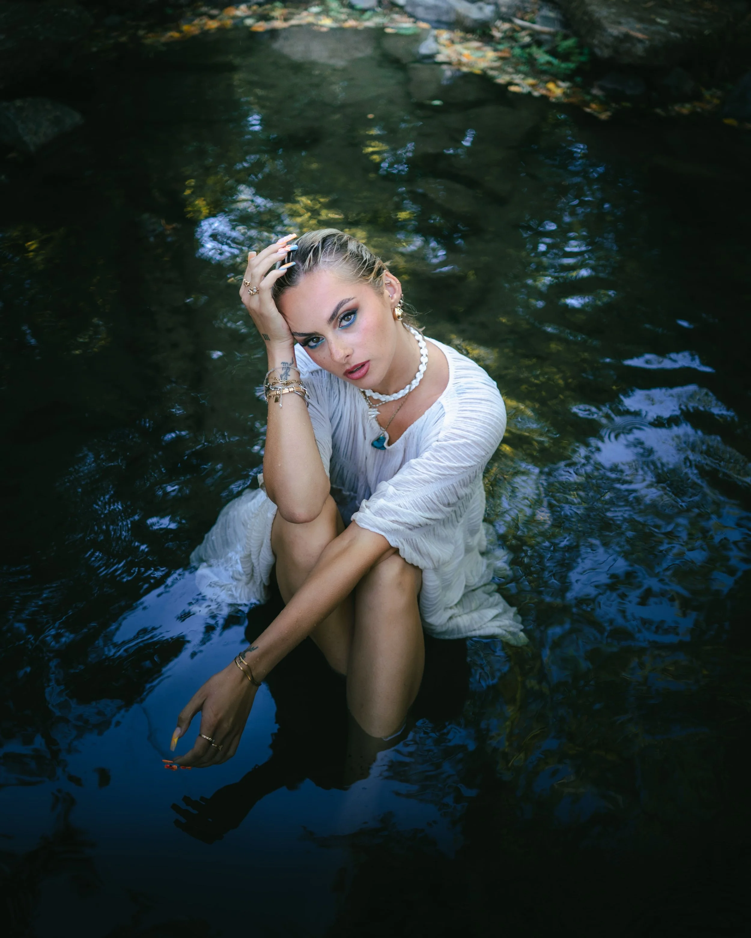 Portrait d'une jeune femme aux cheveux blonds, portant un maquillage prononcé, des bijoux et une robe blanche, est assise dans une rivière, avec ses jambes proches de son corps, en Alsace.