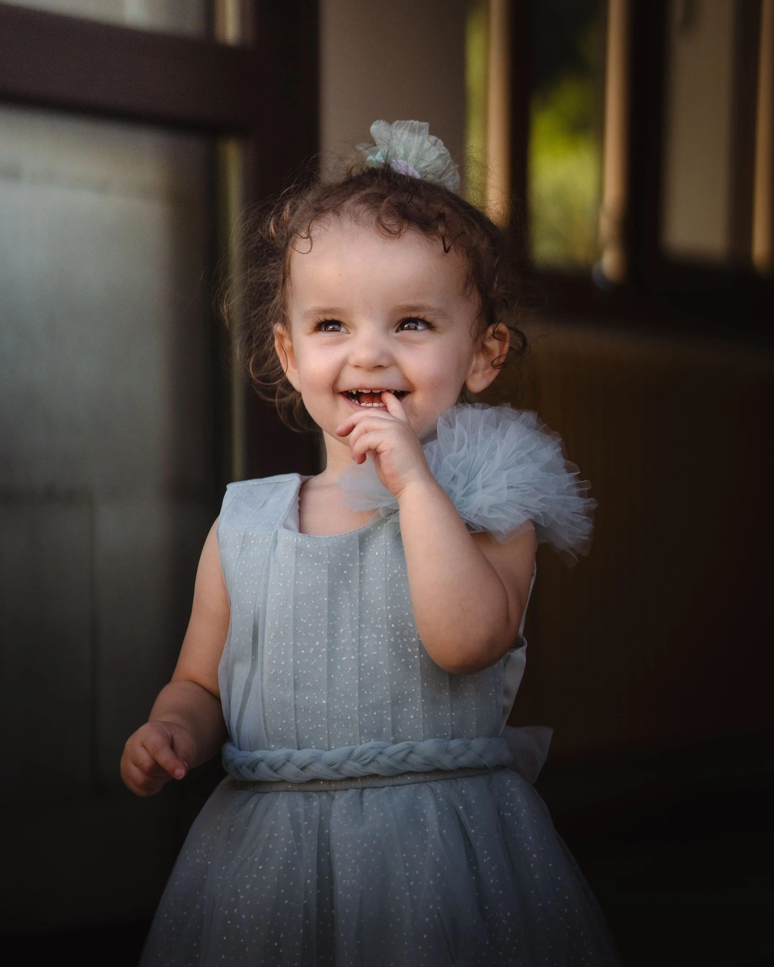 Portrait d'une jeune fille souriante en robe grise avec des tulle, coiffée d'un chignon avec un ruban pastel, dans un intérieur avec rideaux et meubles en bois.