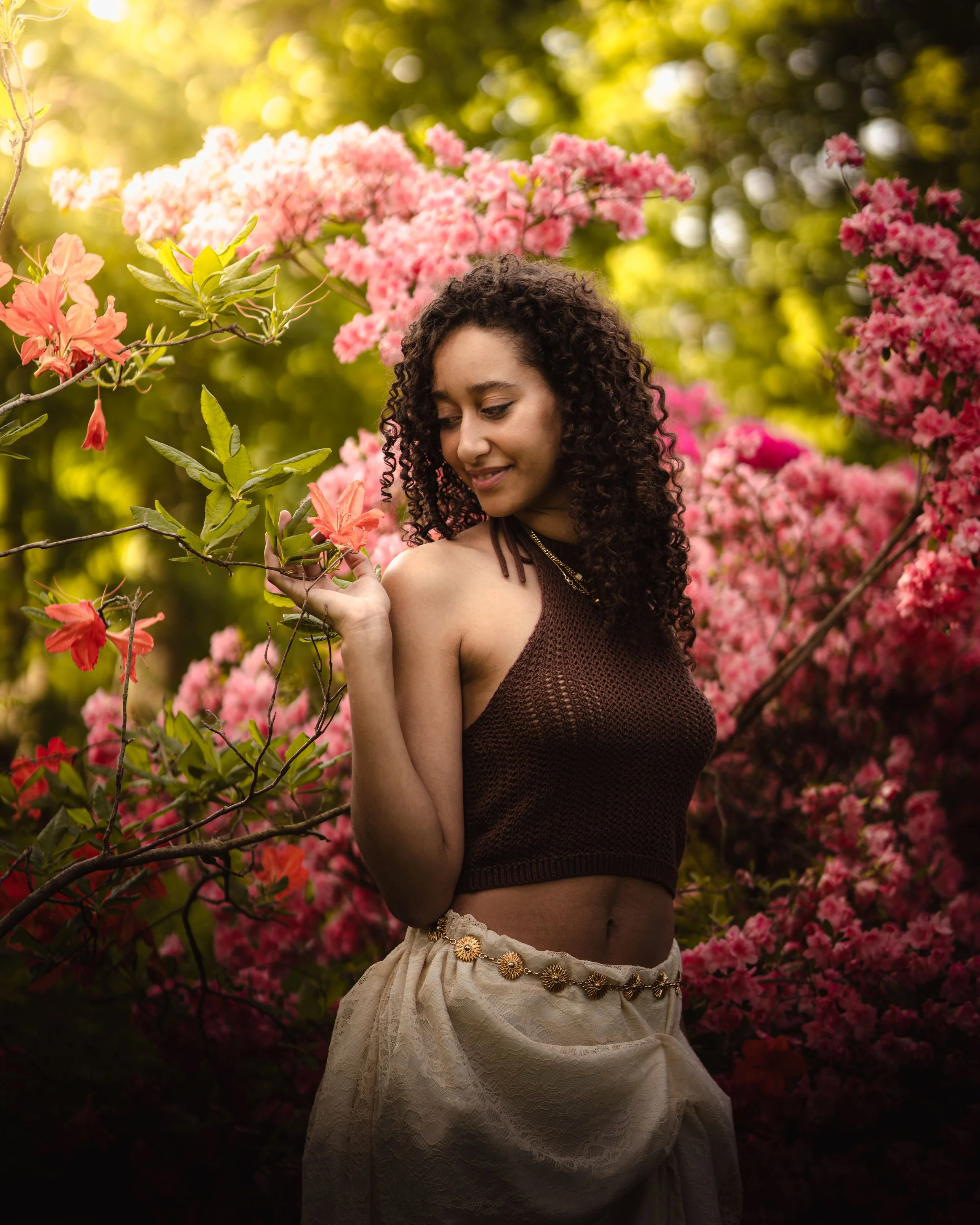 Portrait d'une femme avec des cheveux bouclés, portant une vêtement en filet marron et une jupe longue beige avec une chaîne décorative, se tient parmi des fleurs roses en pleine nature au soleil, au domaine du Windeck.