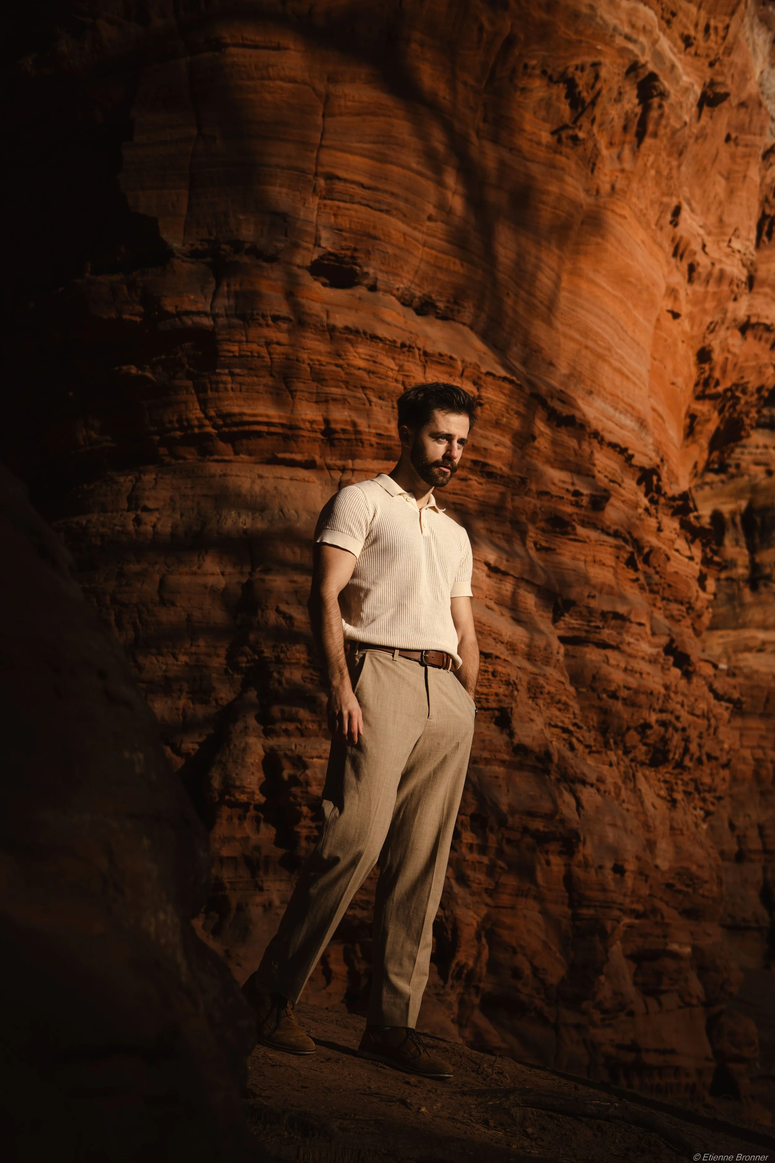 Un homme avec une barbe, portant un polo beige et un pantalon beige, se tient devant une falaise rocheuse rouge du colorado alsacien. Il regarde vers la gauche, dans une posture détendue.