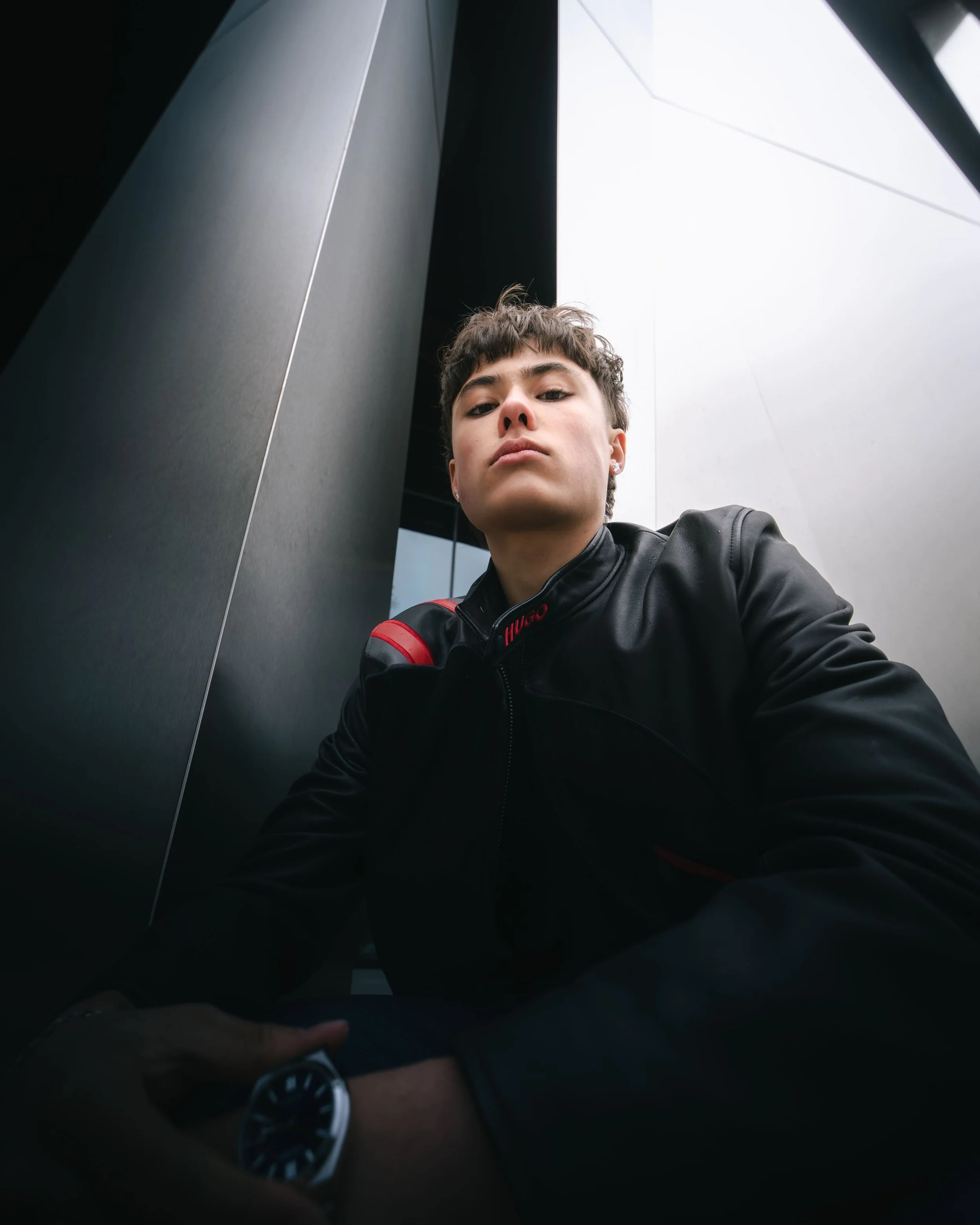 Un jeune homme en veste en cuir noire, regardant vers le haut, assis entre des murs modernes en métal, avec un ciel nuageux en arrière-plan.