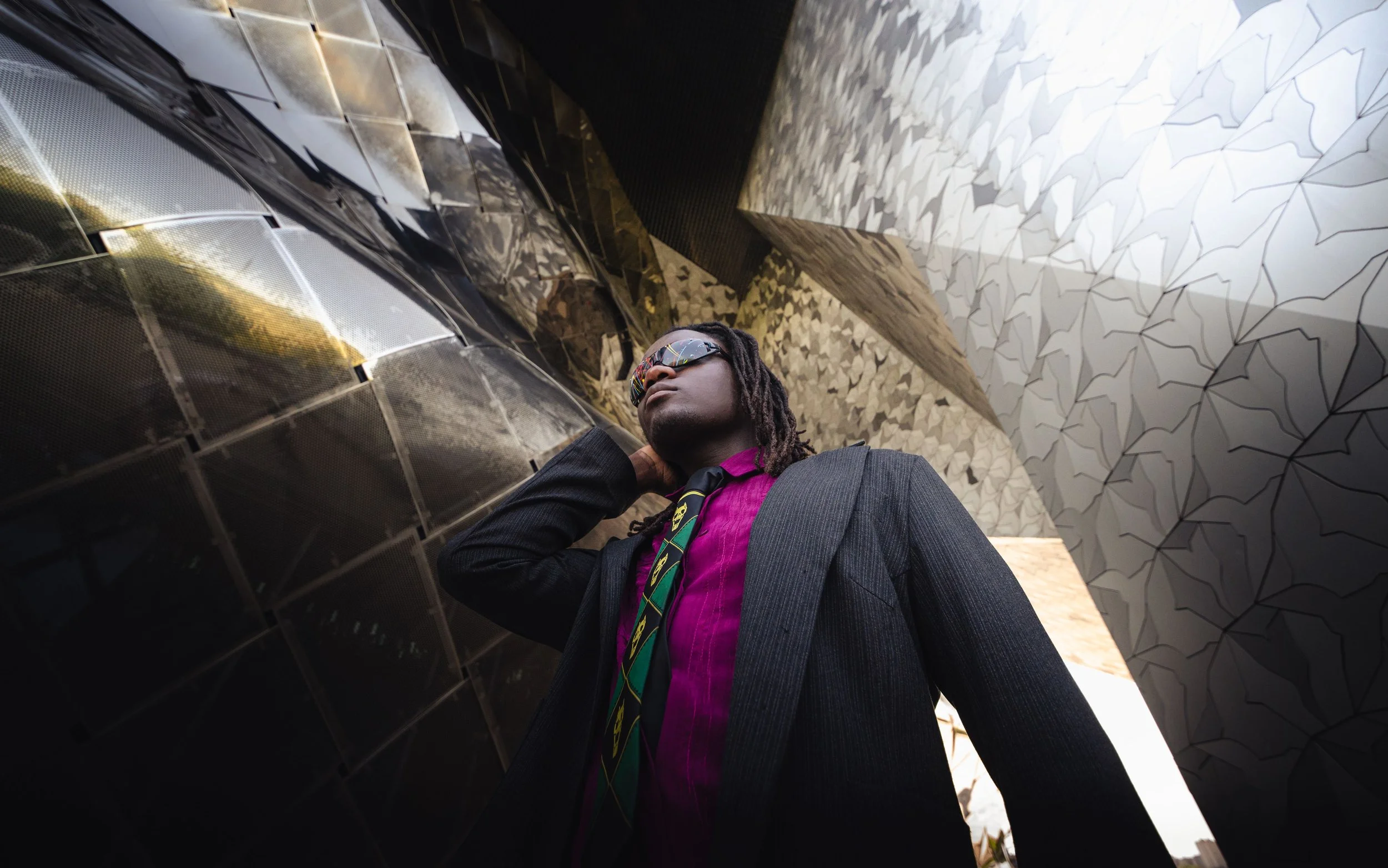 Portrait d'un homme portant des lunettes de soleil, un costume gris, une chemise violette, et une cravate verte avec des motifs, debout sous la structure du philharmonie de Paris.