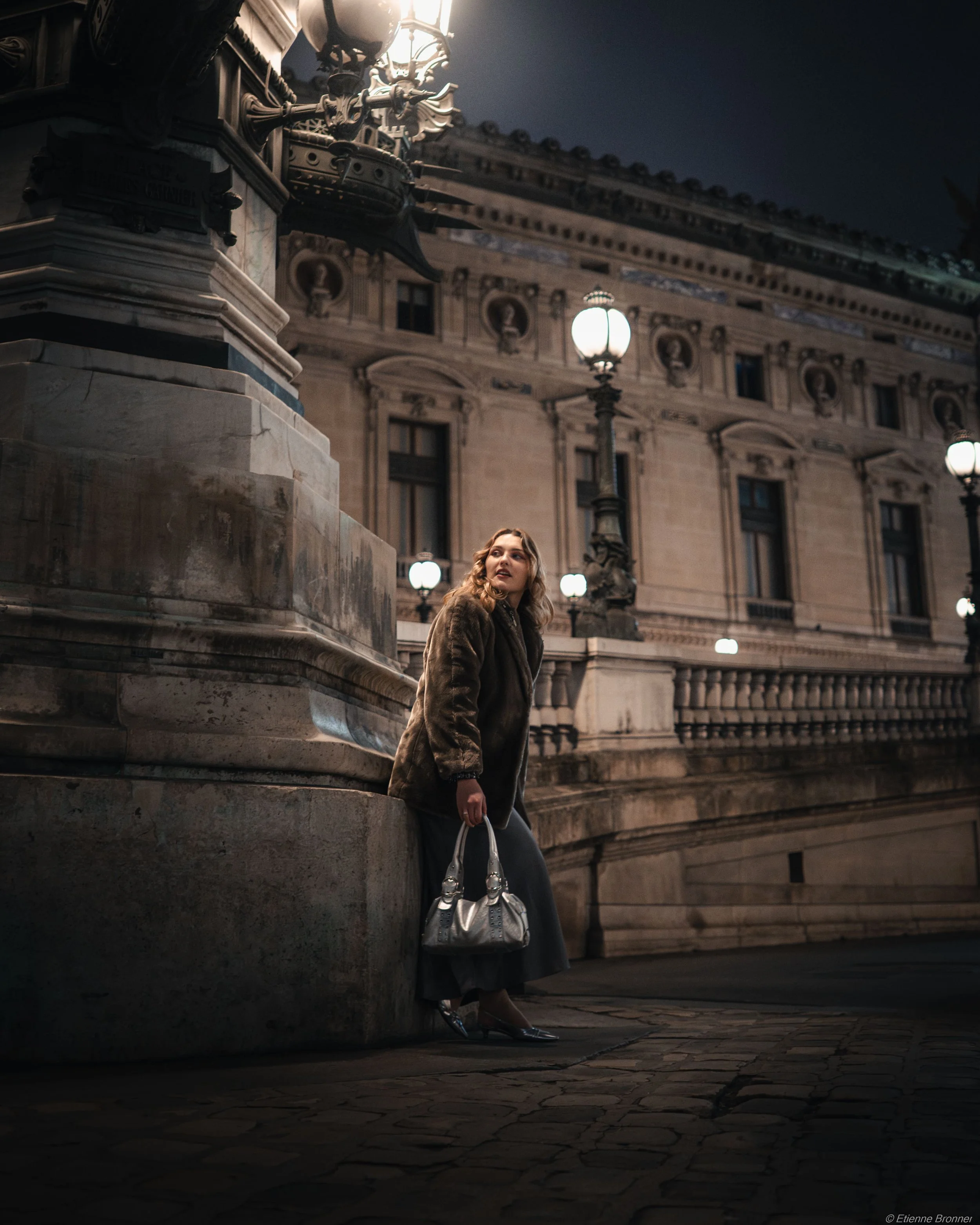 Une femme debout dans la nuit devant un bâtiment historique éclairé par des lampadaires, portant un manteau, une jupe longue, des chaussures à talons et tenant un sac à main métallique.