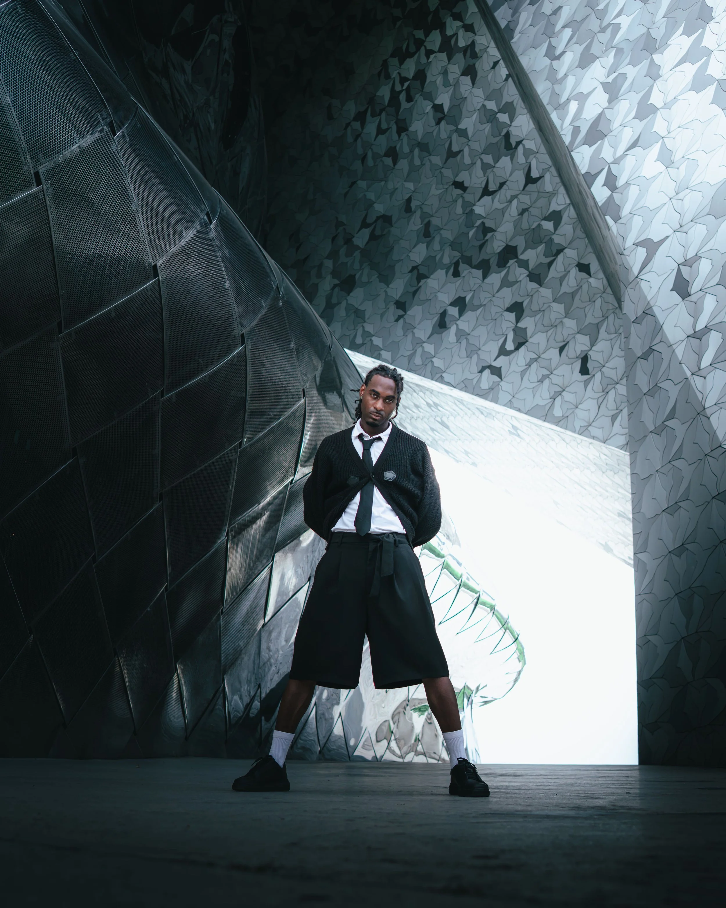 Portrait d'un homme debout sous une structure architecturale moderne en métal et en verre, portant une chemise blanche, une cravate noire, un cardigan noir, une jupe noire, des chaussettes blanches et des baskets noires, au philharmonie de Paris.