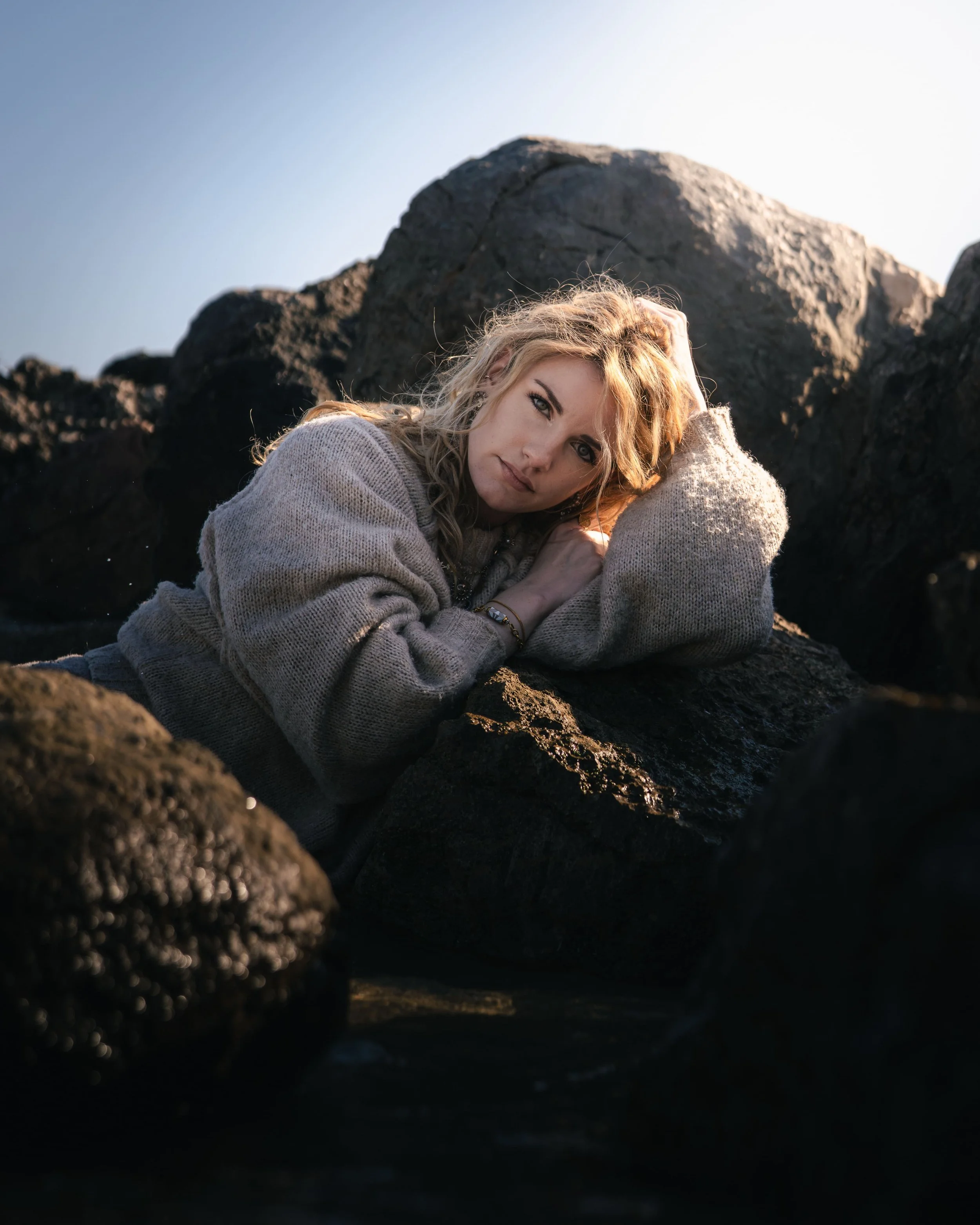 Jeune femme blonde allongée sur des rochers, portant un pull gris, dans un paysage rocheux au soleil levant ou couchant.