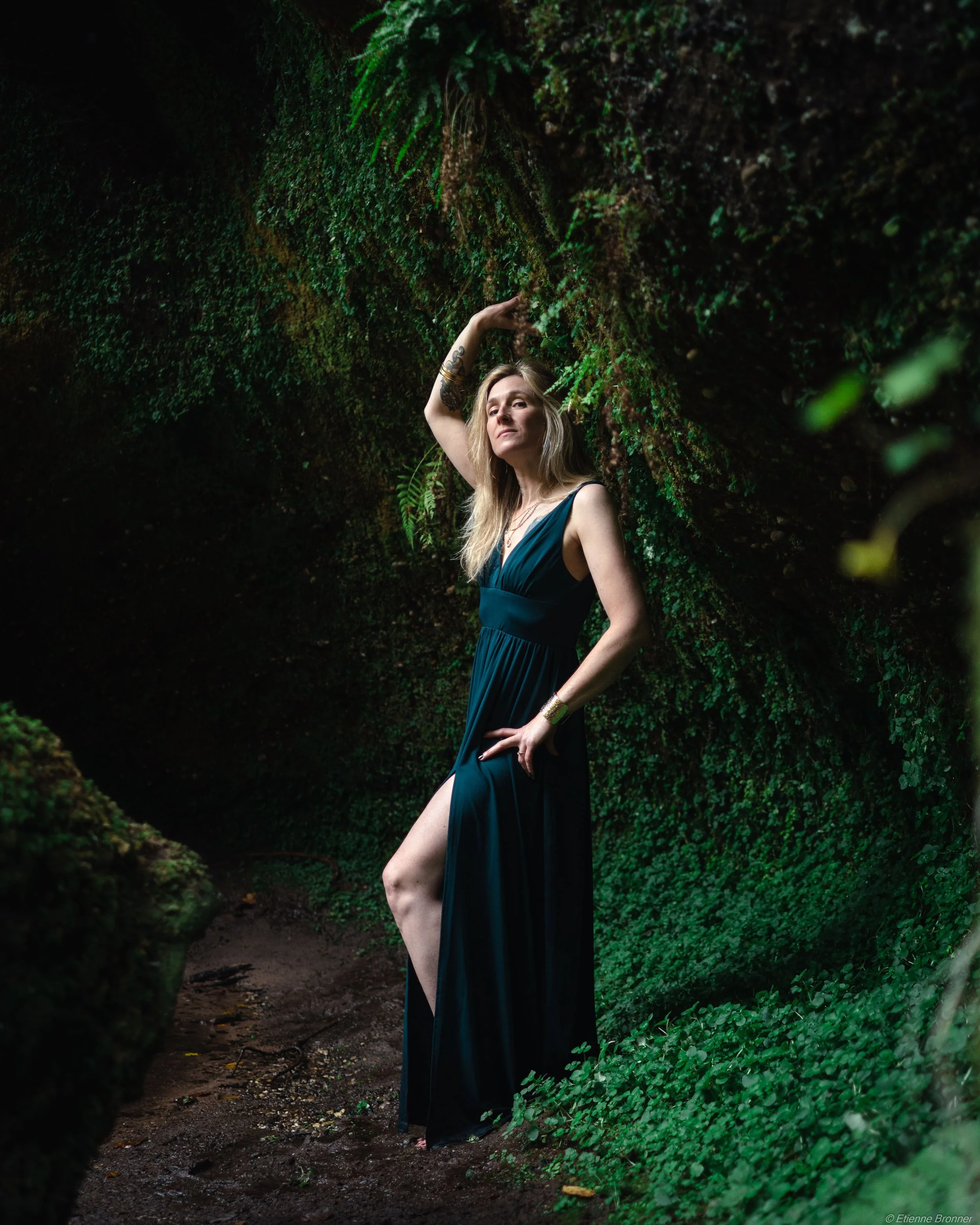 Femme portant une robe longue couleur vert foncé, debout dans une forêt dense avec des feuilles vertes, posant avec une main sur la tête et l'autre sur la poitrine, ambiance mystérieuse.