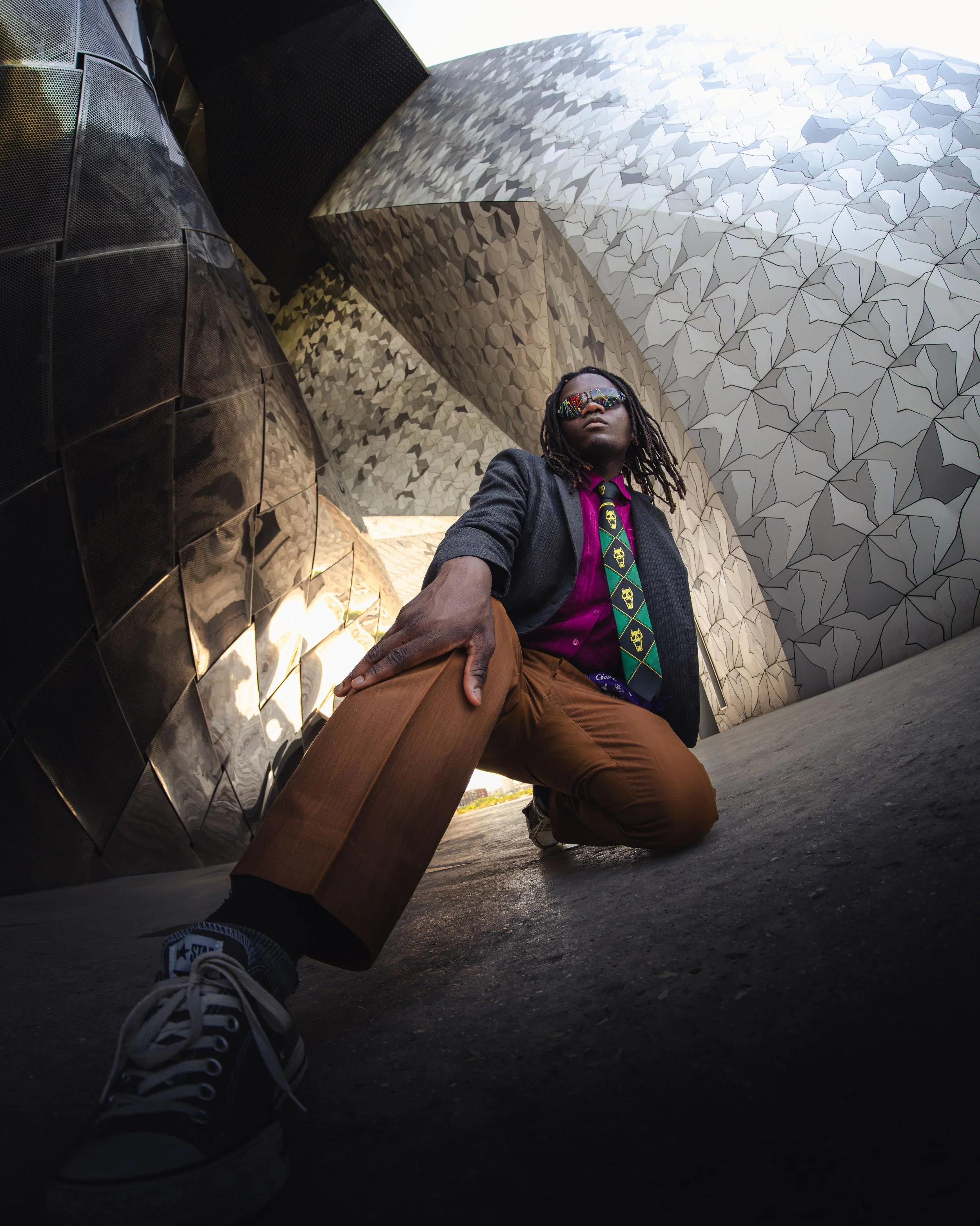 Portrait d'un homme avec des dreadlocks portant des lunettes de soleil colorées, une veste noire, une chemise violette et une cravate, posé à côté d'un bâtiment architectural moderne au philharmonie de Paris.