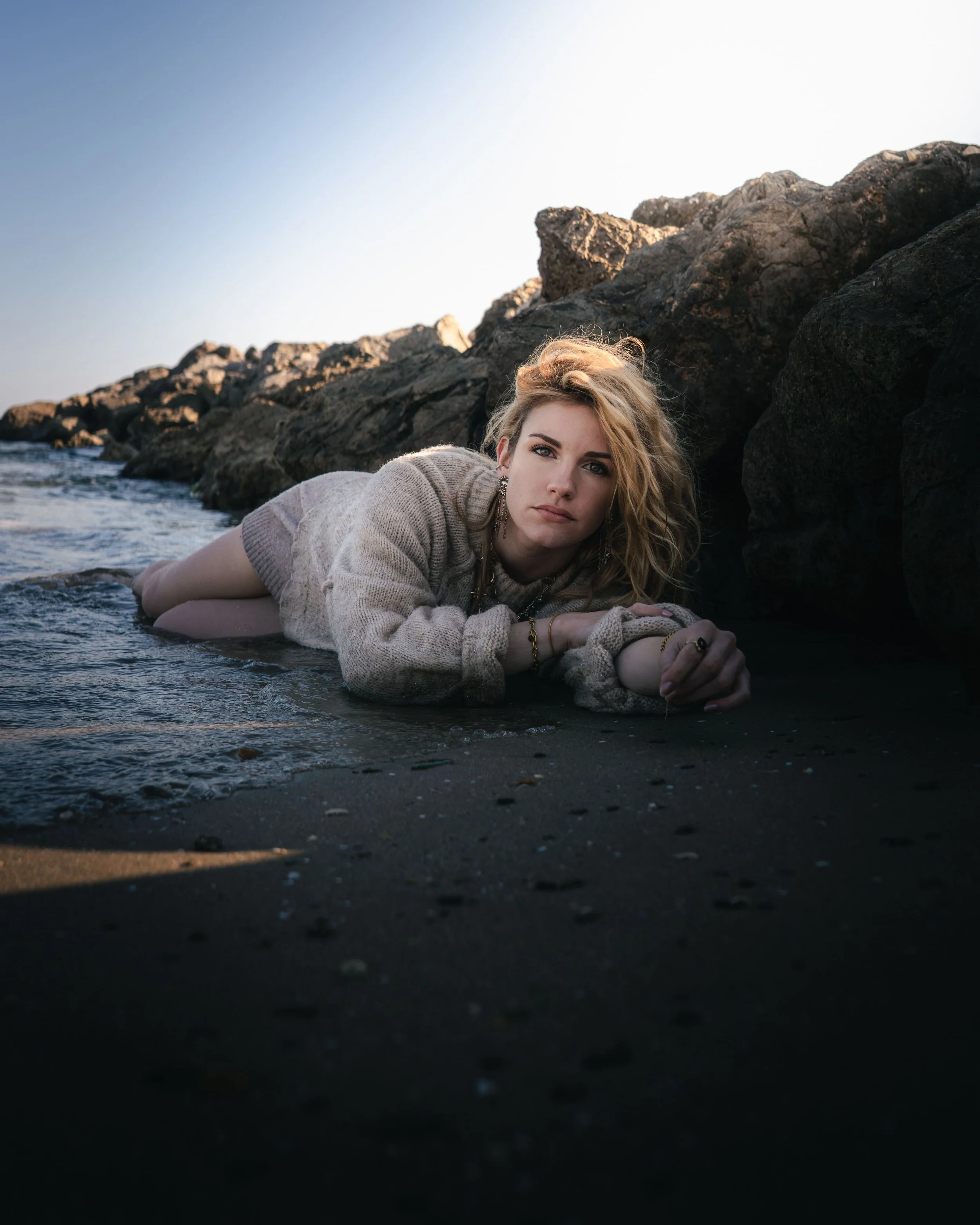 Jeune femme allongée sur la plage près des rochers, regardant la caméra, portant un pull beige et des bijoux.