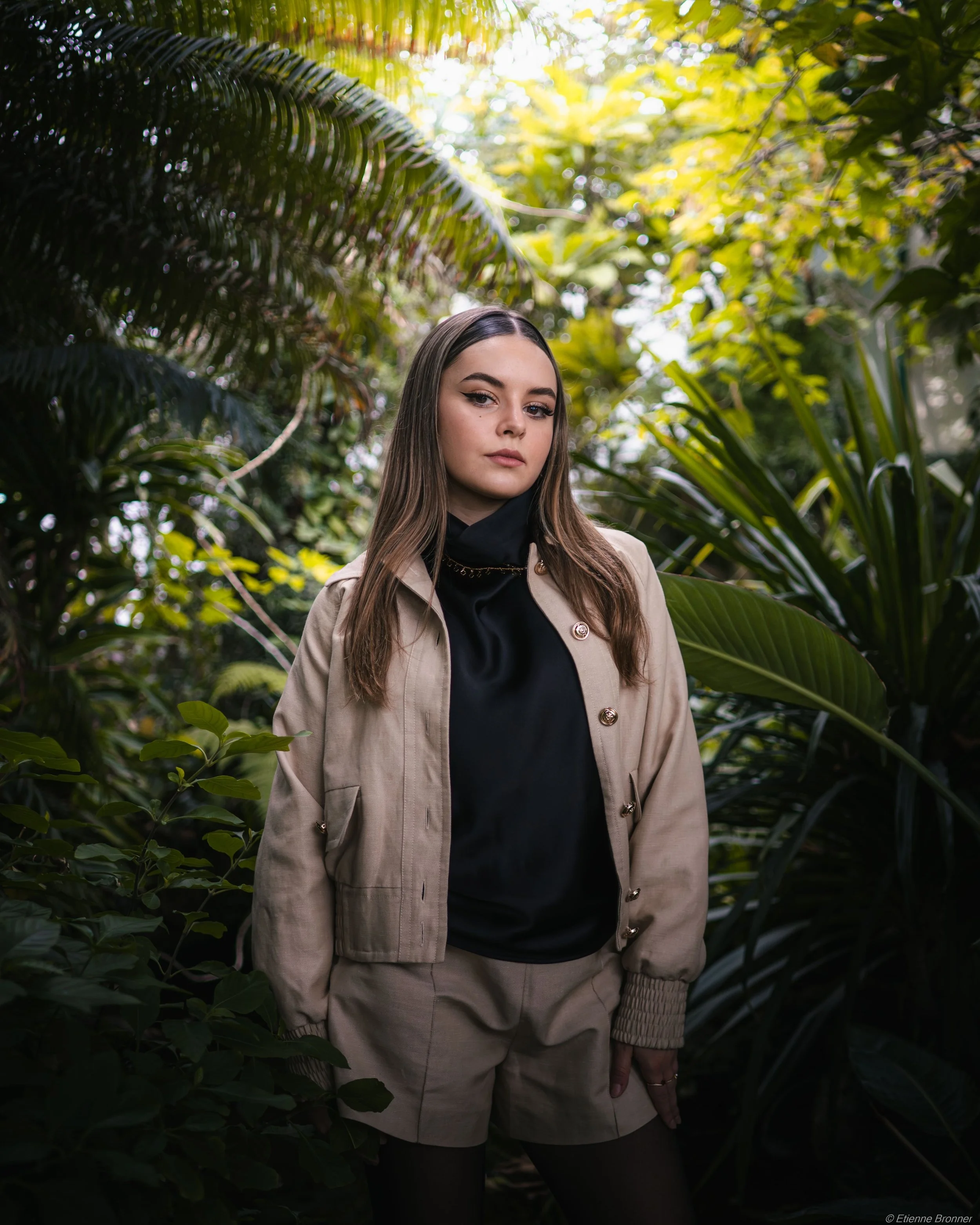 Portrait d'une jeune femme vêtue d'une veste beige et d'un haut noir, posant parmi les plantes tropicales des serres d'Auteuil à Paris.
