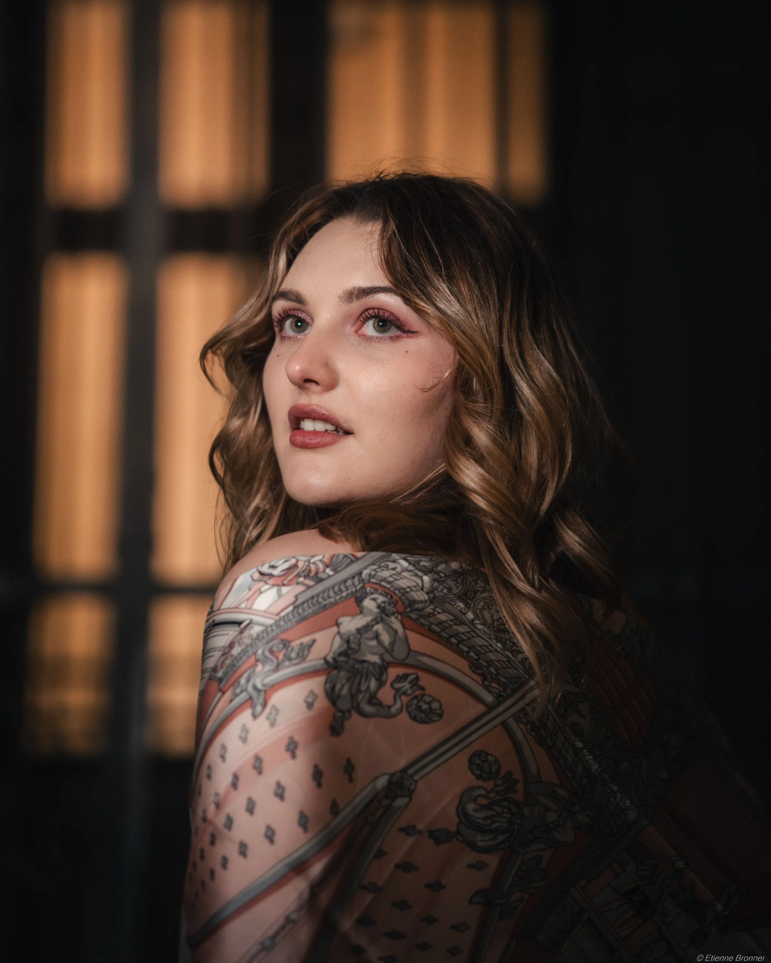 Portrait d'une jeune femme avec des cheveux bruns bouclés, portant une robe avec un motif complexe, dans un environnement sombre avec une lumière chaude en arrière-plan.