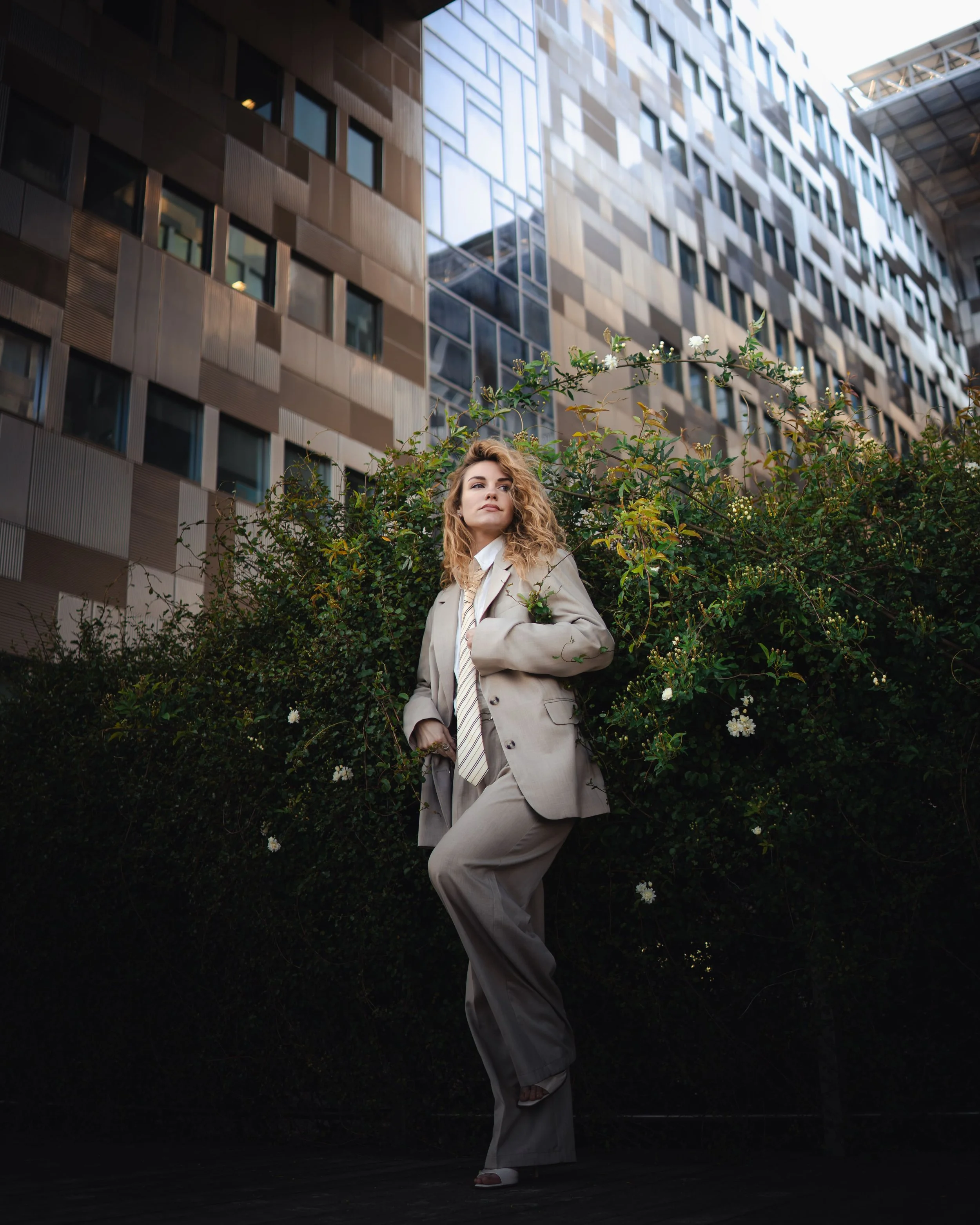 Une femme en costume beige debout devant une haie avec un bâtiment moderne en arrière-plan.