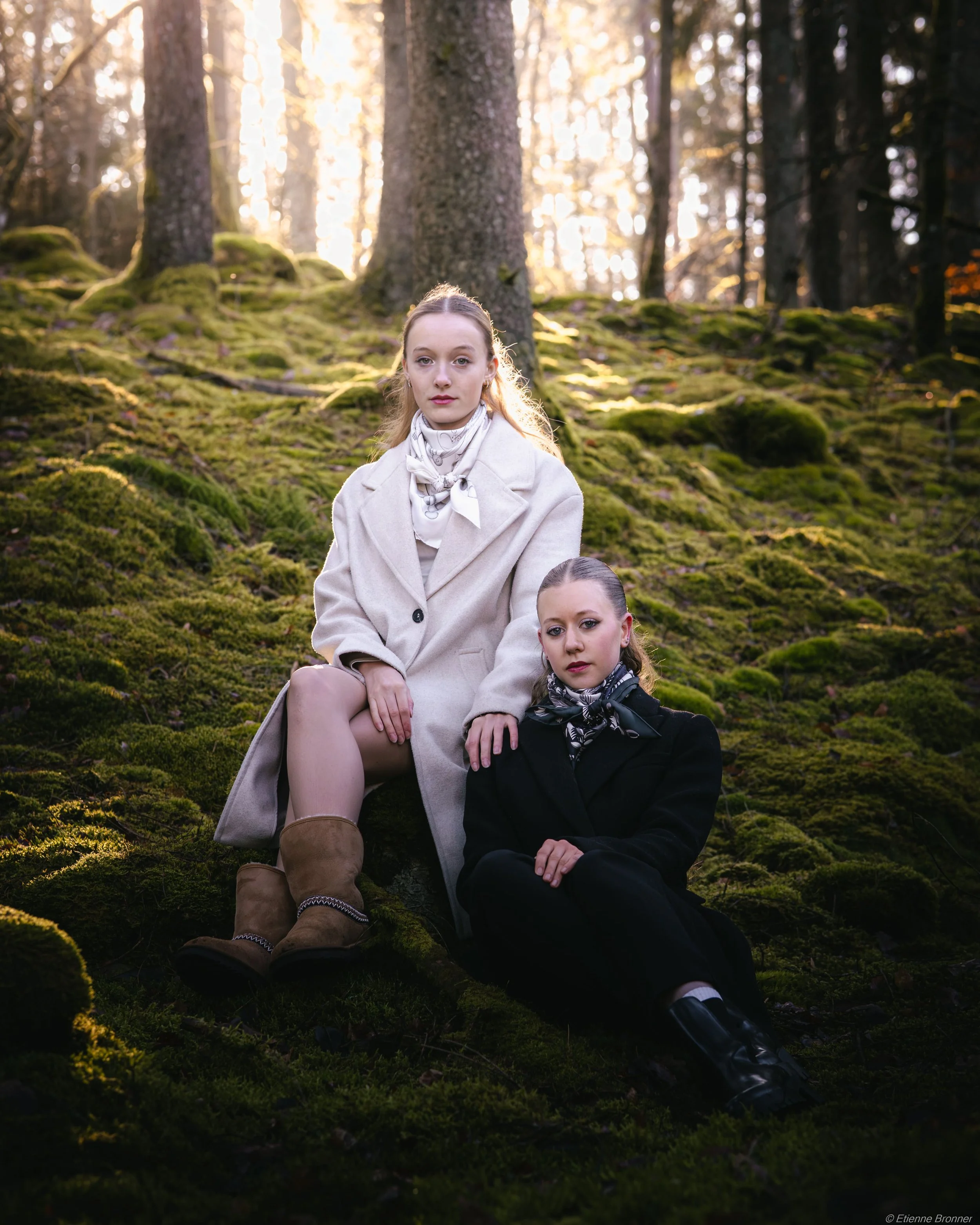 Portrait de deux jeunes femmes assises dans une forêt des Vosges avec des arbres et de la mousse au sol, éclairées par la lumière du soleil qui filtre à travers les arbres.