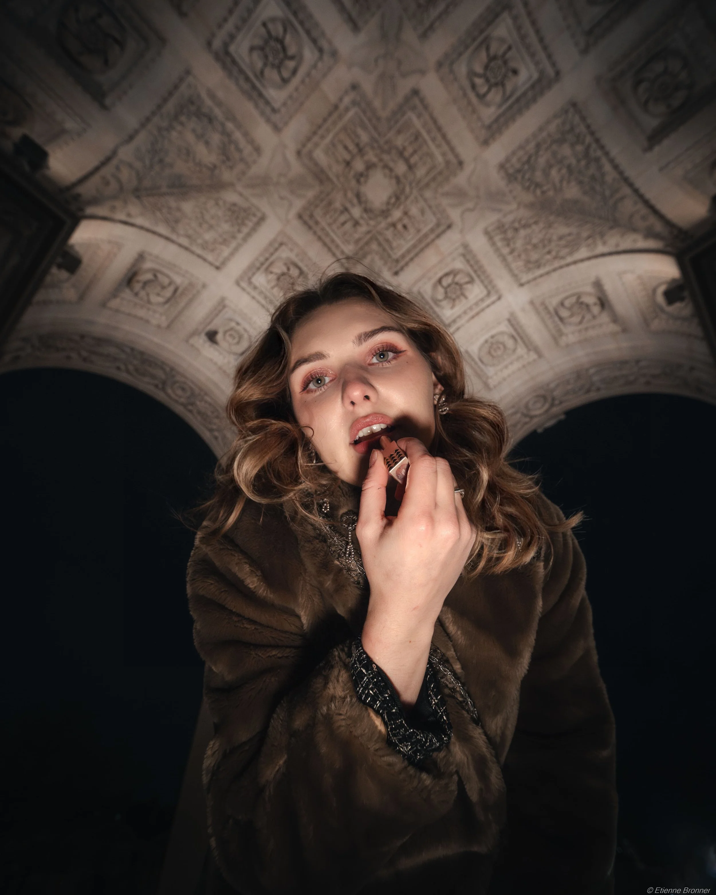 Une jeune femme aux cheveux bouclés portant un manteau en fausse fourrure foncé, appliquant du maquillage sur ses lèvres dans une pièce avec un plafond orné de motifs géométriques du l'arche du Louvre.