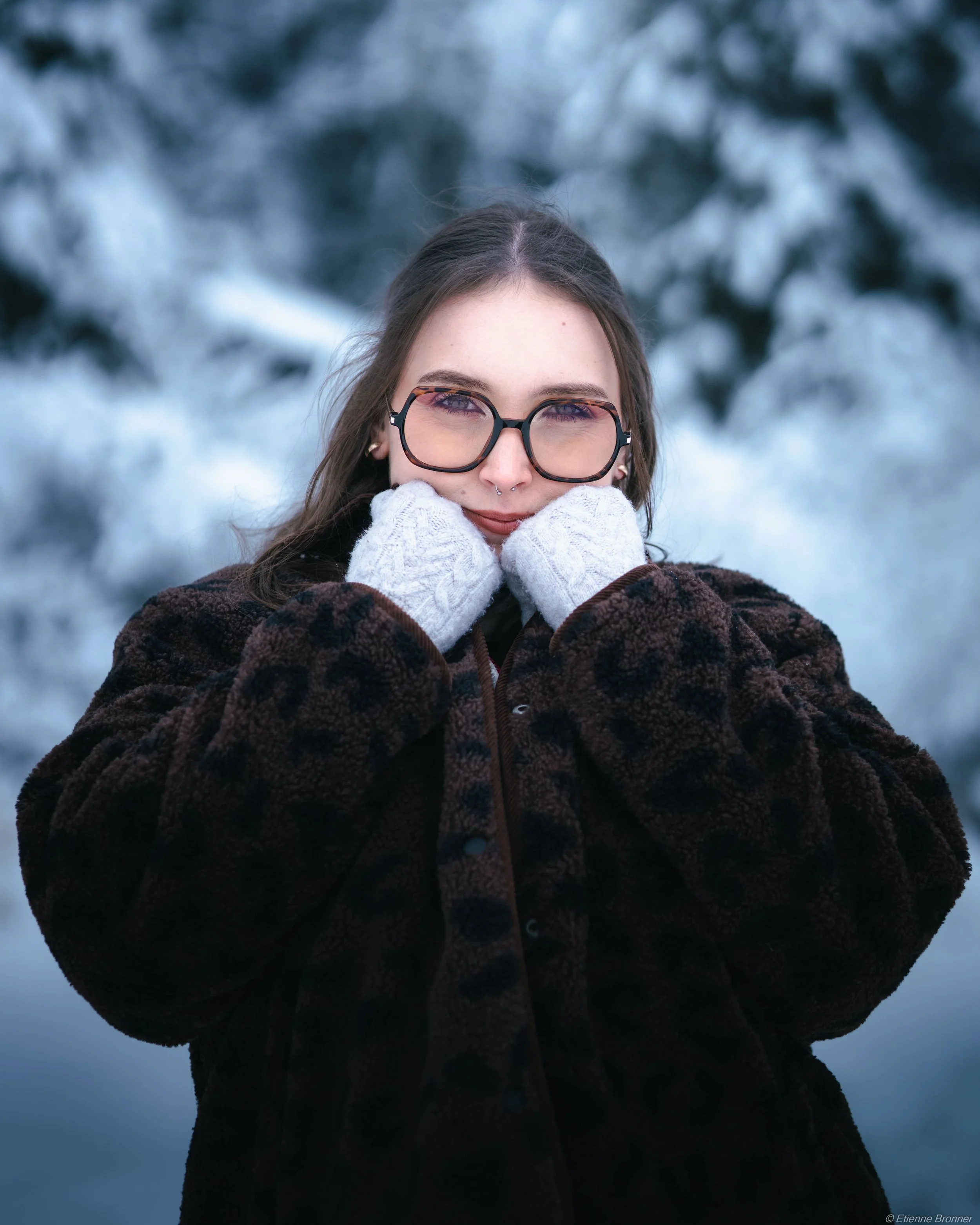 Portrait d'une jeune femme en manteau marron et lunettes, tenant son visage avec des gants blancs, dans un décor enneigé.