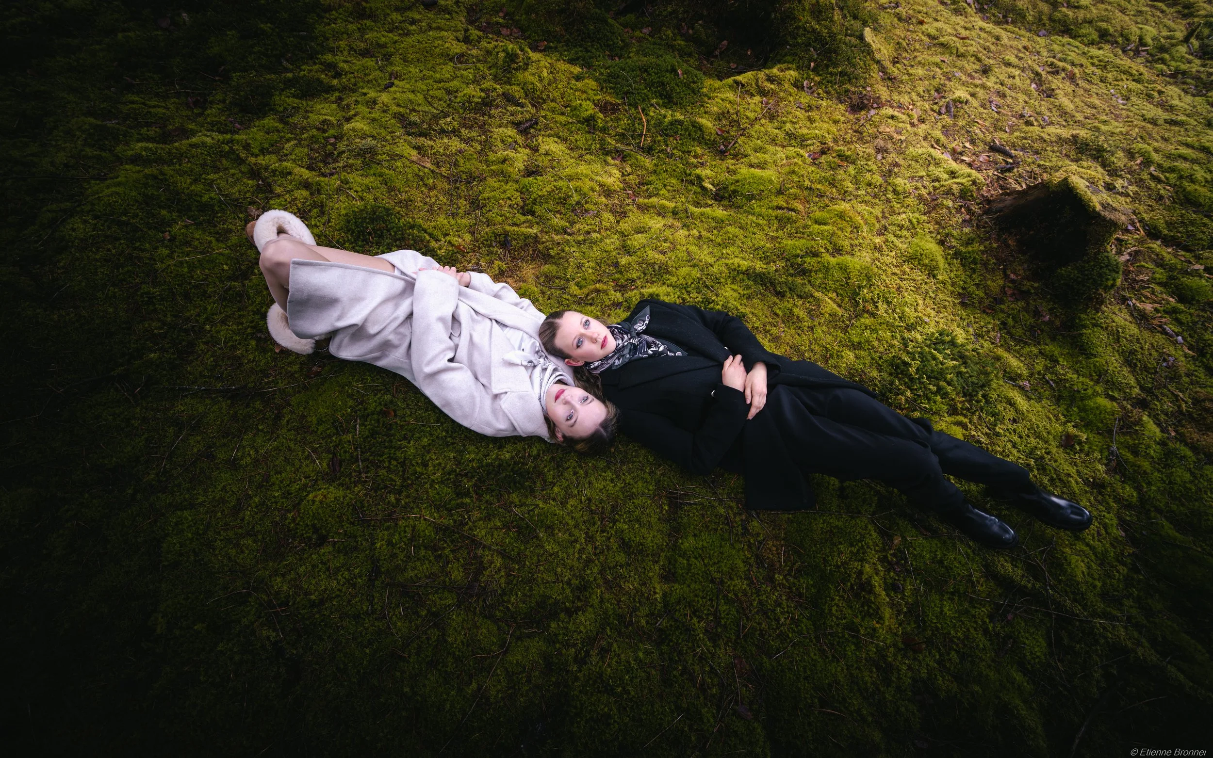 Portrait de deux femmes allongées sur un tapis de mousse verte dans une forêt, regardant le haut de l'appareil photo, l'une en manteau blanc et l'autre en costume noir.
