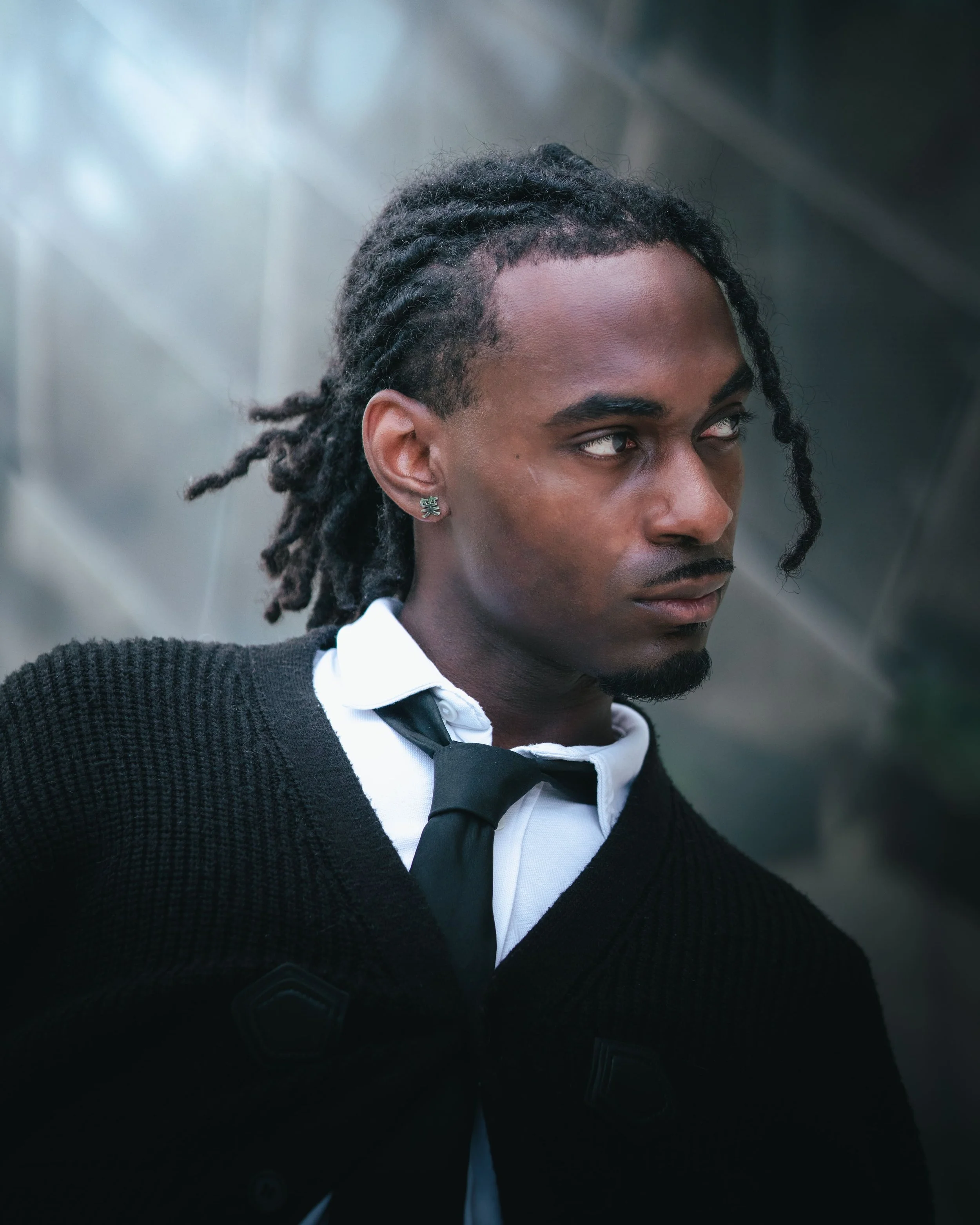 Portrait d'un homme aux dreadlocks, portant une chemise blanche, une cravate noire, un pull noir et une boucle d'oreille en forme de caractère chinois, au philharmonie de Paris