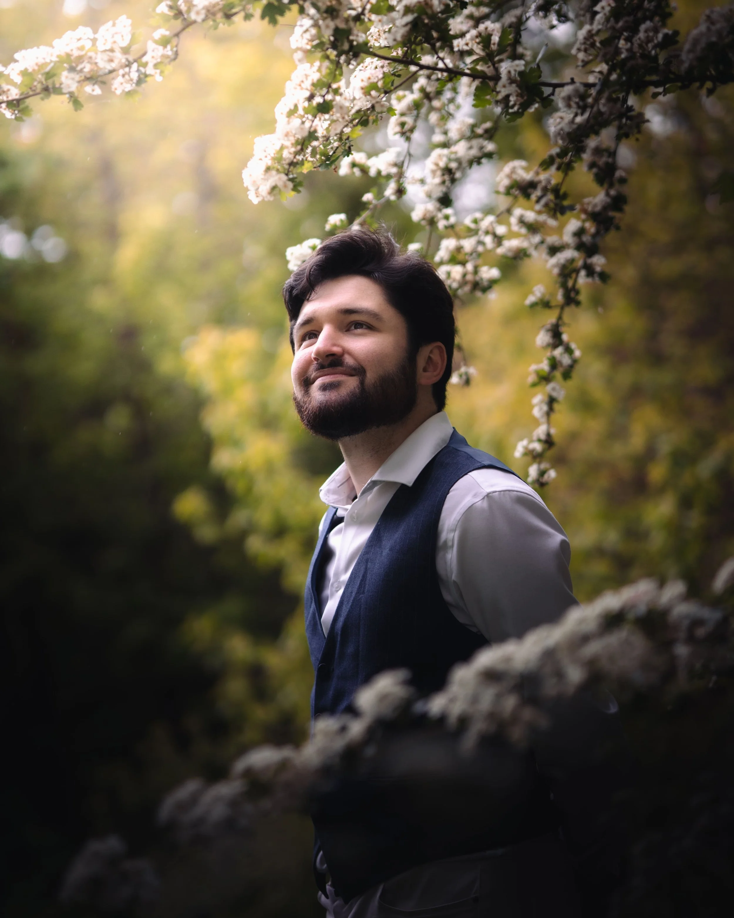 Portrait d'un homme avec une barbe porte un costume formel sur une chemise blanche, se tenant devant un arbre en fleurs dans un parc ou un jardin pendant la journée, à Nantes.