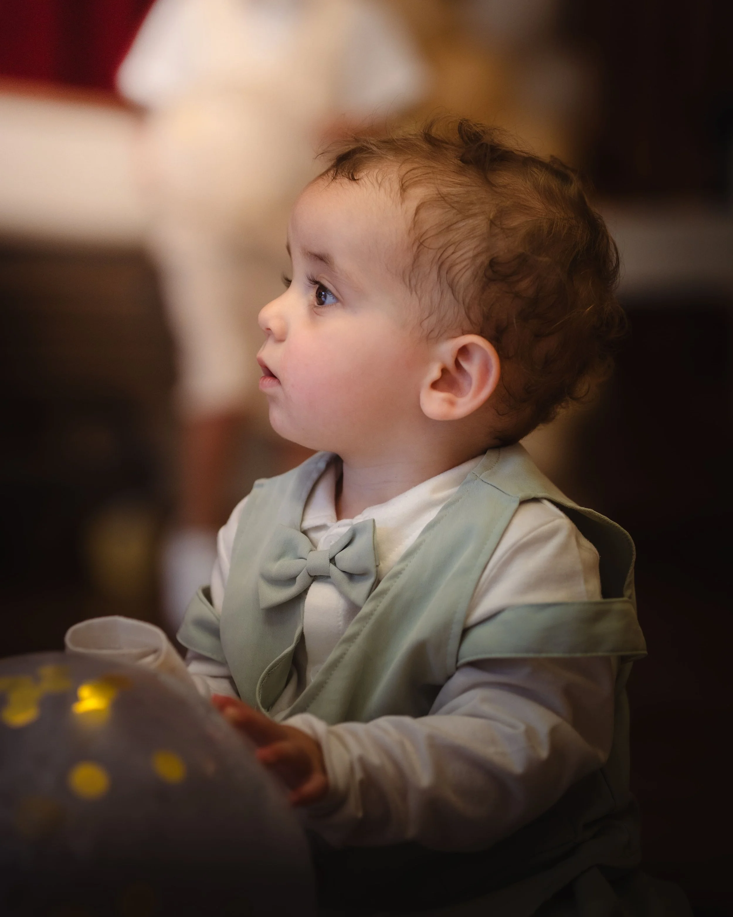 Portrait d'un jeune garçon avec des cheveux bouclés, vu de profil, portant une veste beige avec un nœud papillon, assis à une table, dans un environnement intérieur.