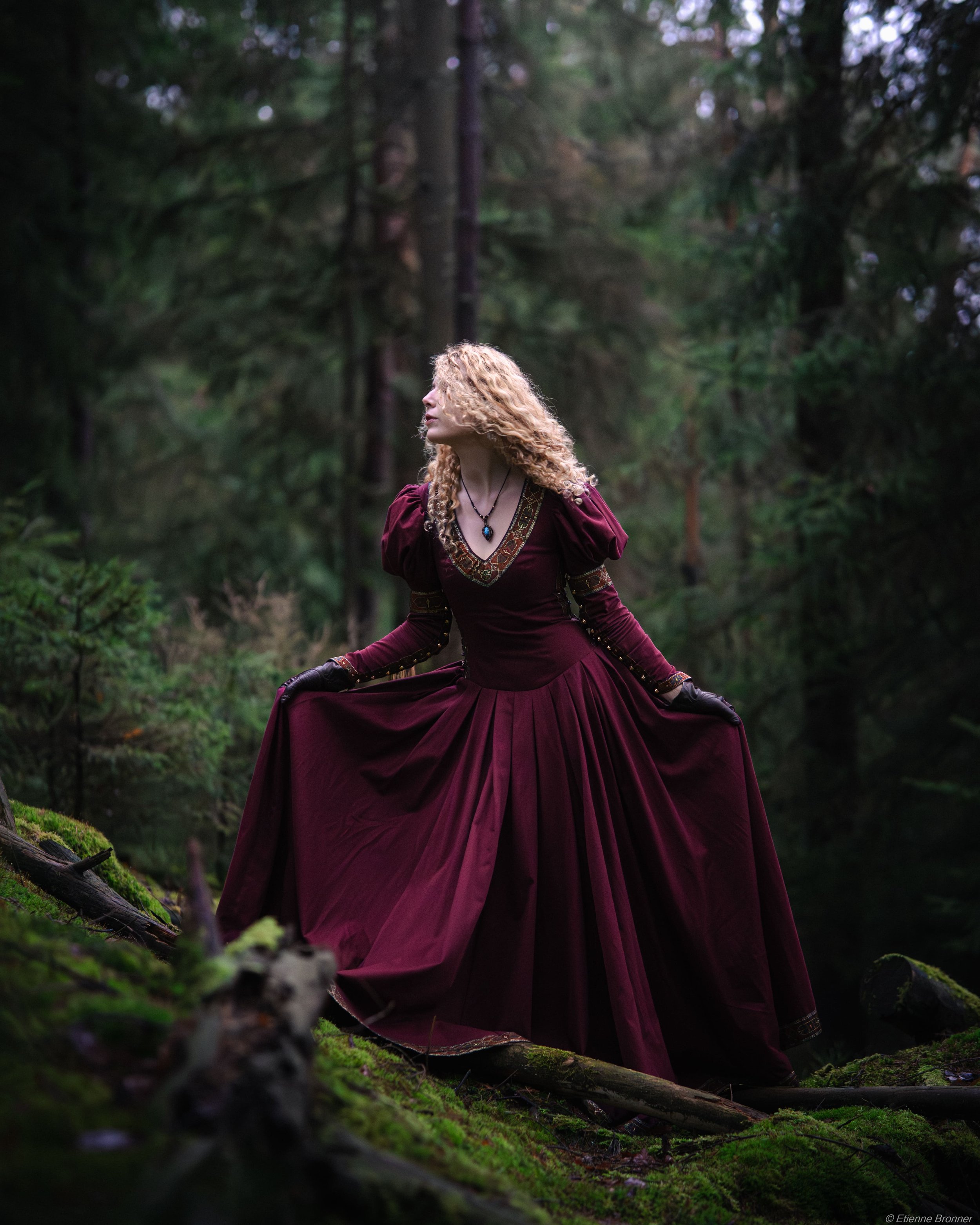 Portrait d'une femme portant une longue robe de couleur Bordeaux dans une forêt des Vosges verdoyante, tenant le bas de sa robe. Elle a des cheveux bouclés blonds et porte des bijoux noirs, avec des gants noirs, dans une scène de nature mystérieuse.