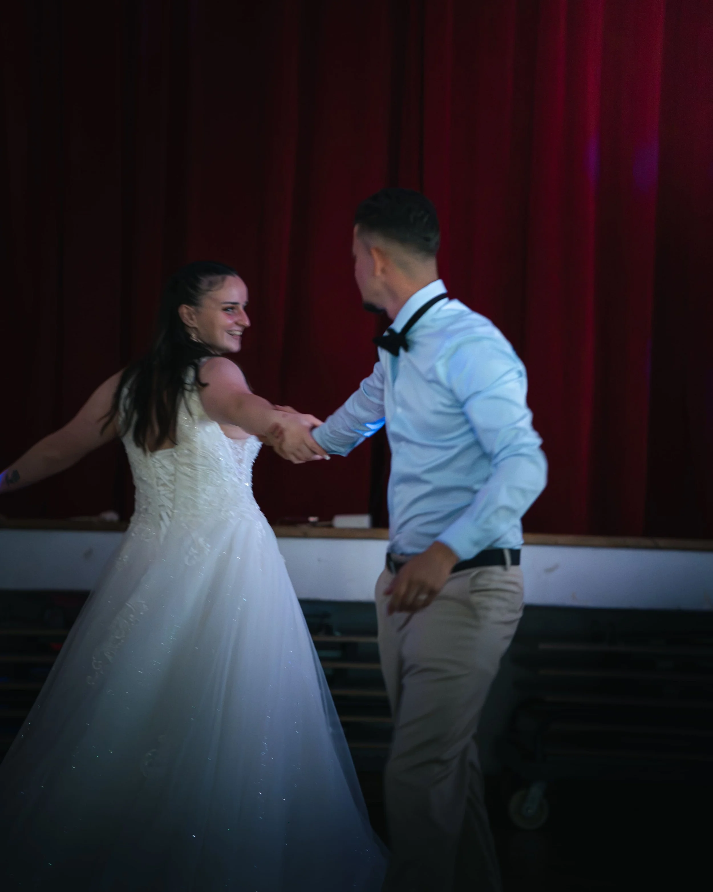 Un couple danse lors d'une cérémonie, avec la femme en robe de mariée et le homme en costume blanc, dans une salle avec rideaux rouges en arrière-plan.