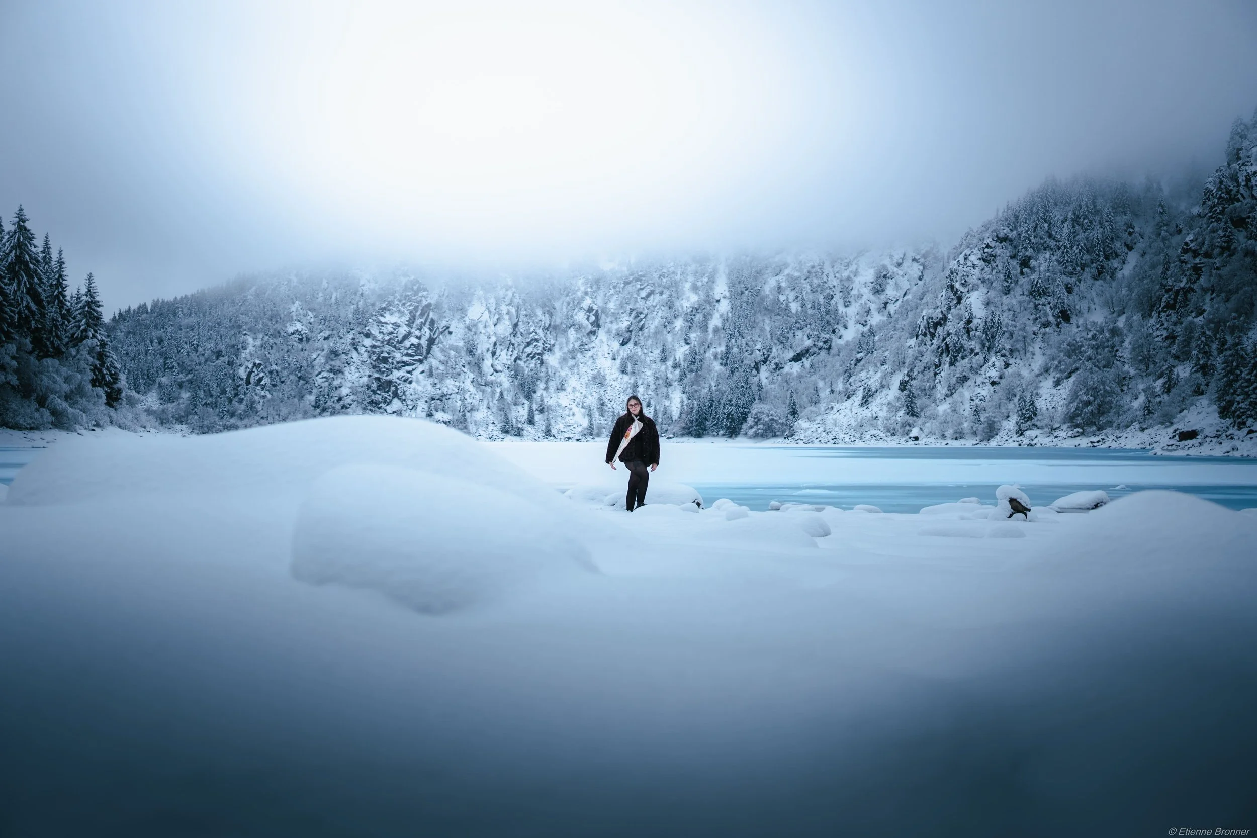 Portrait devant le lac blanc d'une personne qui marche dans une vaste étendue de neige près d'un lac gelé, entourée par une forêt enneigée sous un ciel nuageux.