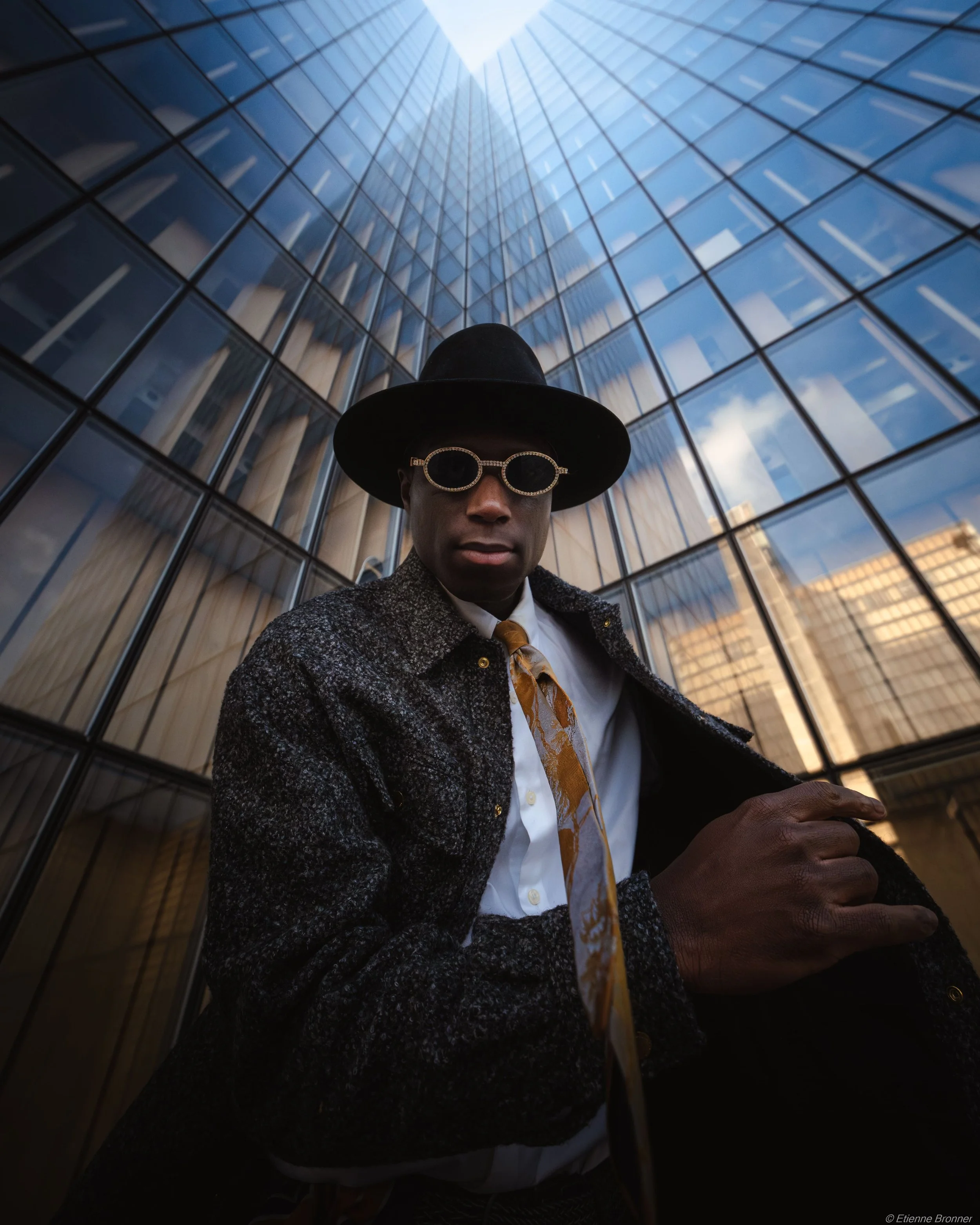 Un homme portant des lunettes de soleil, un chapeau noire et un manteau sombre, debout devant un bâtiment en verre moderne dans une ville.
