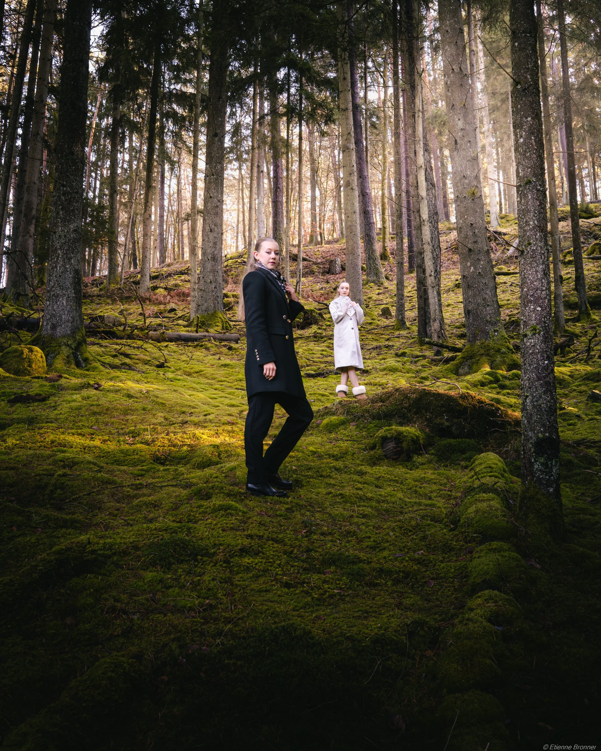 Portrait de deux femmes debout dans une forêt des Vosges, l'une en manteau noir et l'autre en manteau blanc, entourées d'arbres et recouvertes de mousse.