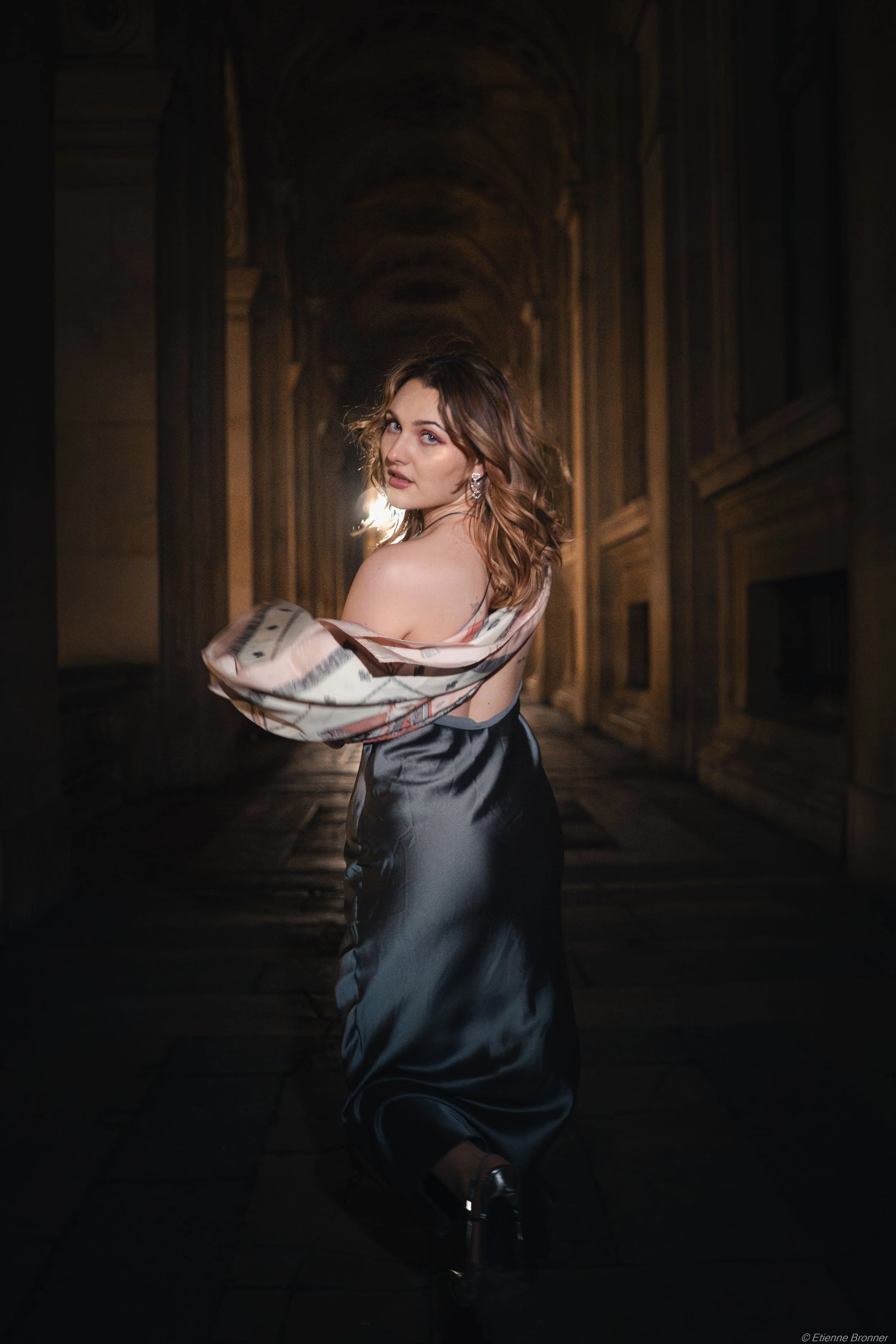 Une femme habillée en robe de soirée satinée dans un couloir historique en pierre, éclairée en contre-jour.