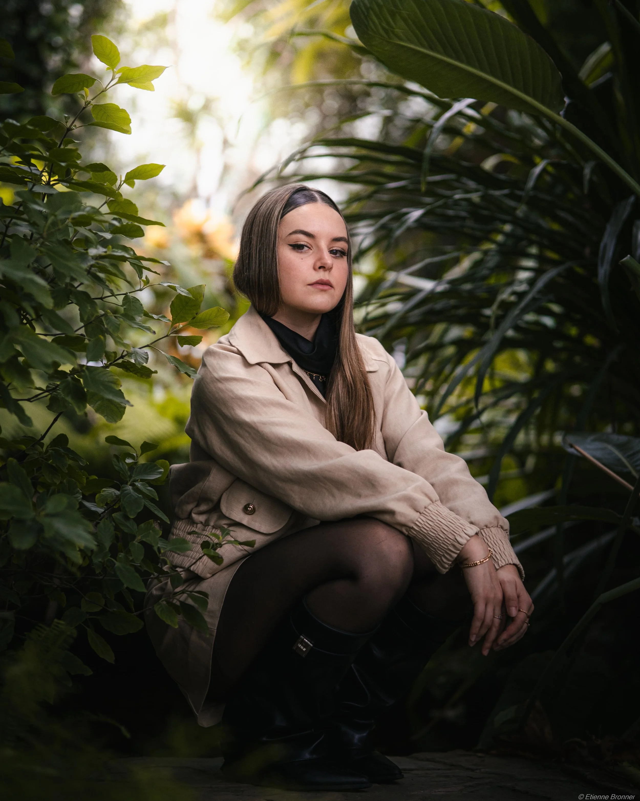 Jeune femme assise parmi la végétation dense des serres d'Auteuil dans un environnement naturel, portant un manteau beige, des collants noirs et des bottes en cuir noir, avec une expression neutre.