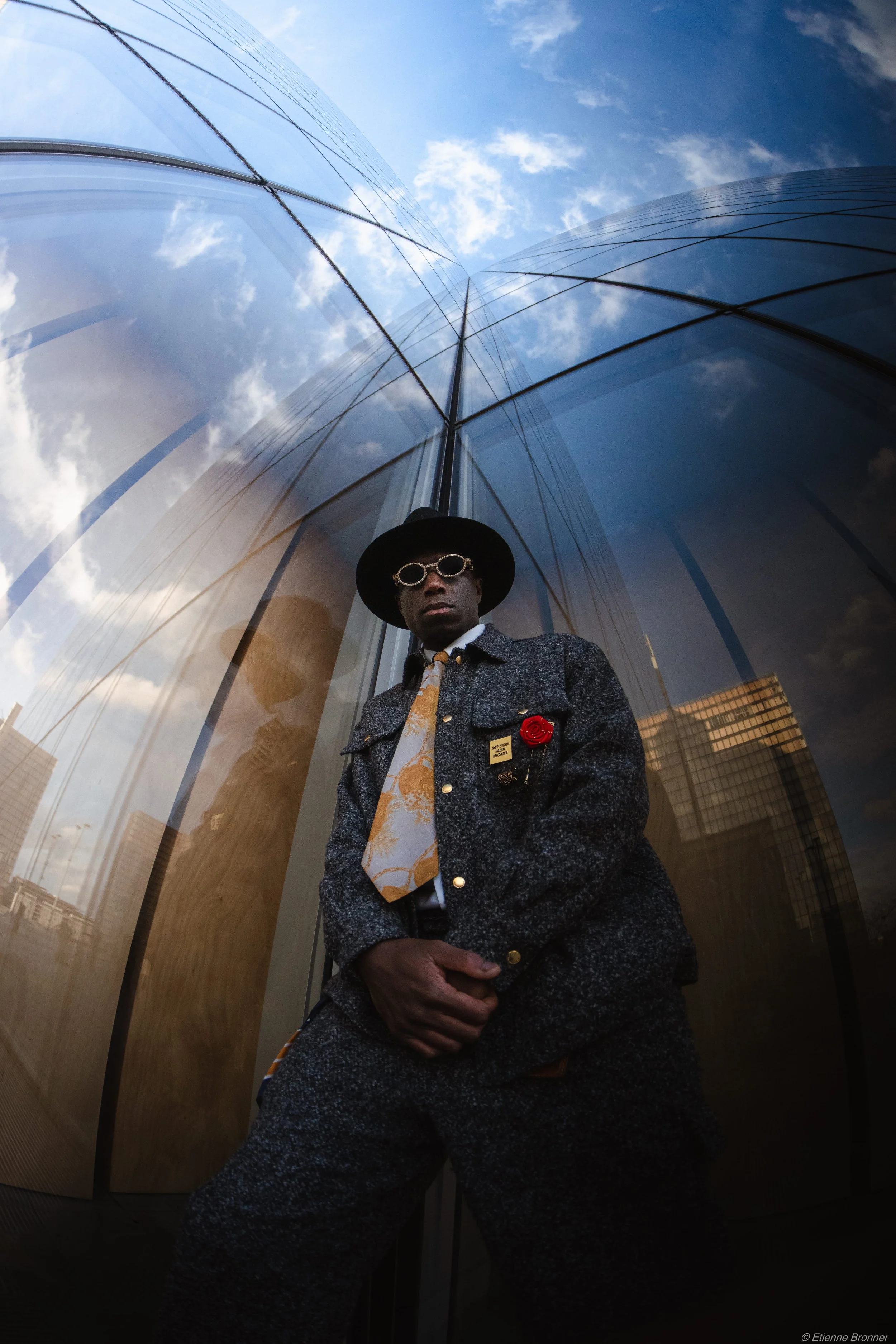 Un homme portant des lunettes de soleil, un chapeau sombre, une veste en tweed, une cravate avec un motif floral, et une broche en forme de rose rouge, posé devant un bâtiment en verre avec des reflets du ciel et des immeubles de la BNF de Paris.