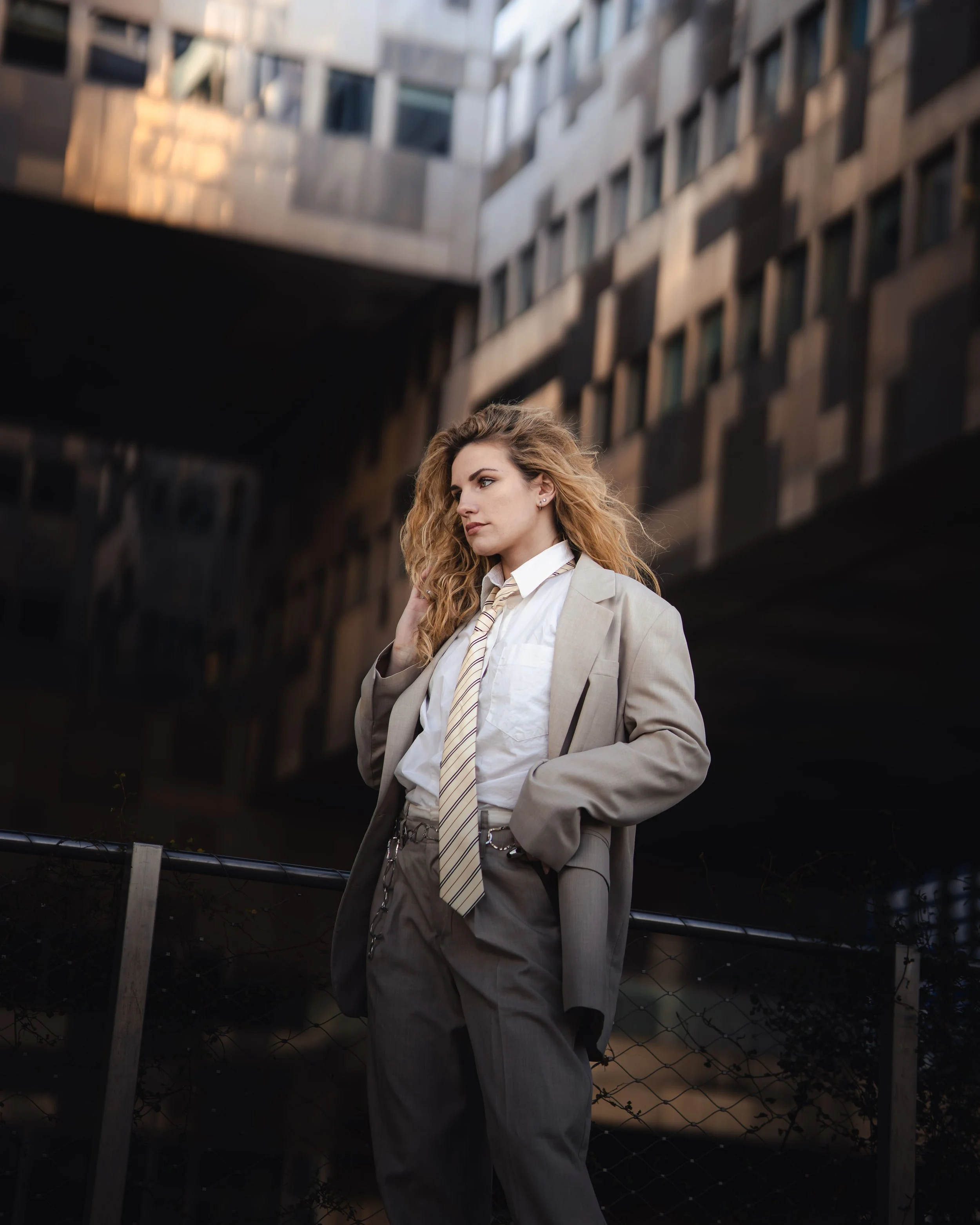 Une femme aux cheveux bouclés portant un costume beige et une cravate dans un environnement urbain moderne.