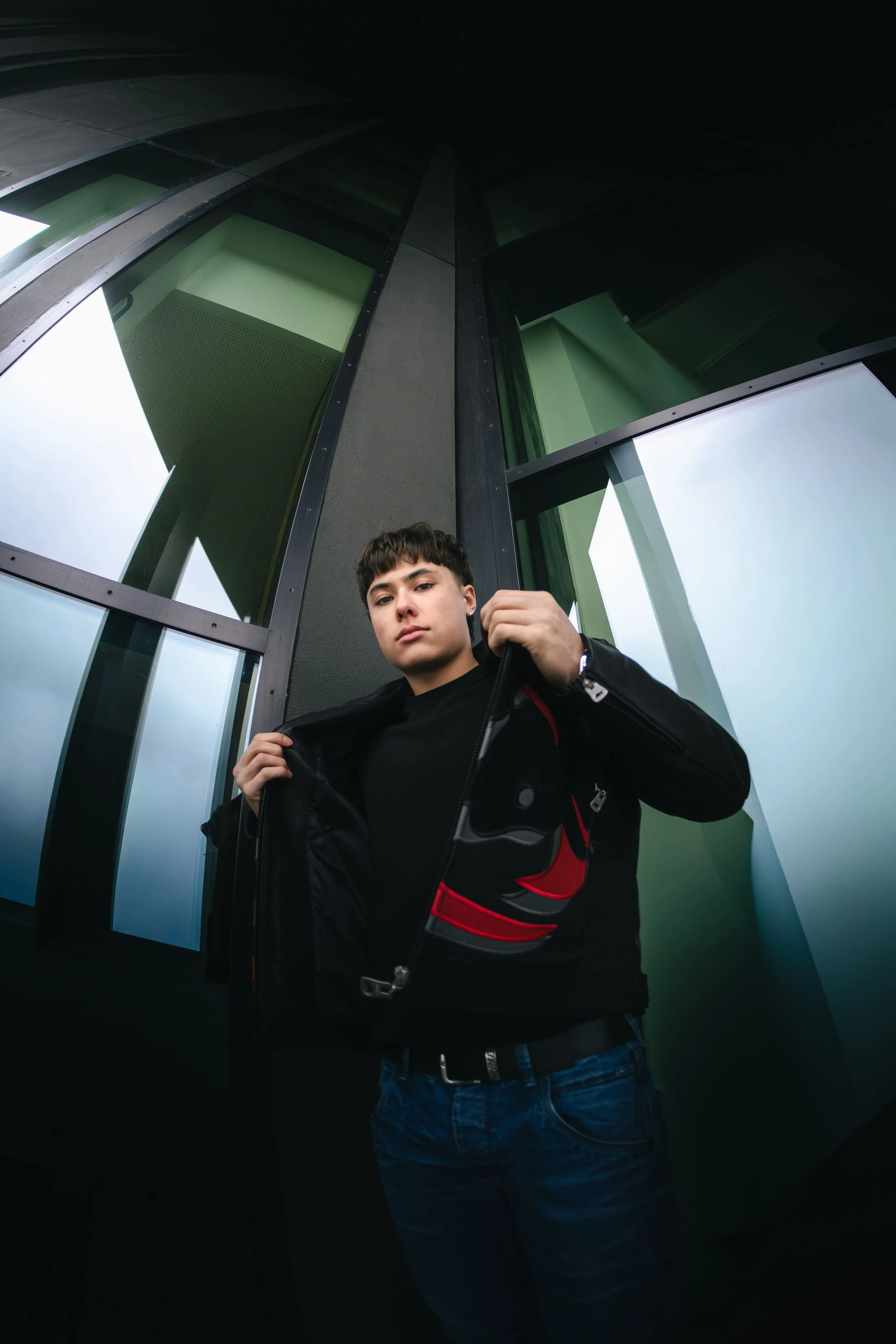Jeune homme debout devant un bâtiment moderne en verre, portant une veste noire et une ceinture noire, regardant vers la caméra.