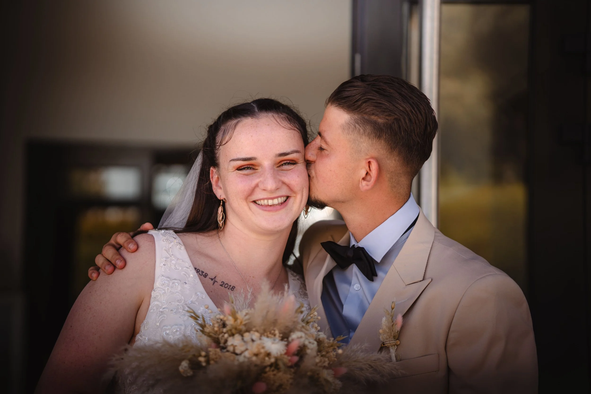 Portrait d'un couple de mariés souriants, la femme portant une robe de mariée blanche et le homme en costume beige avec une chemise bleue et un noeud papillon noir, lors de leur mariage en Alsace.